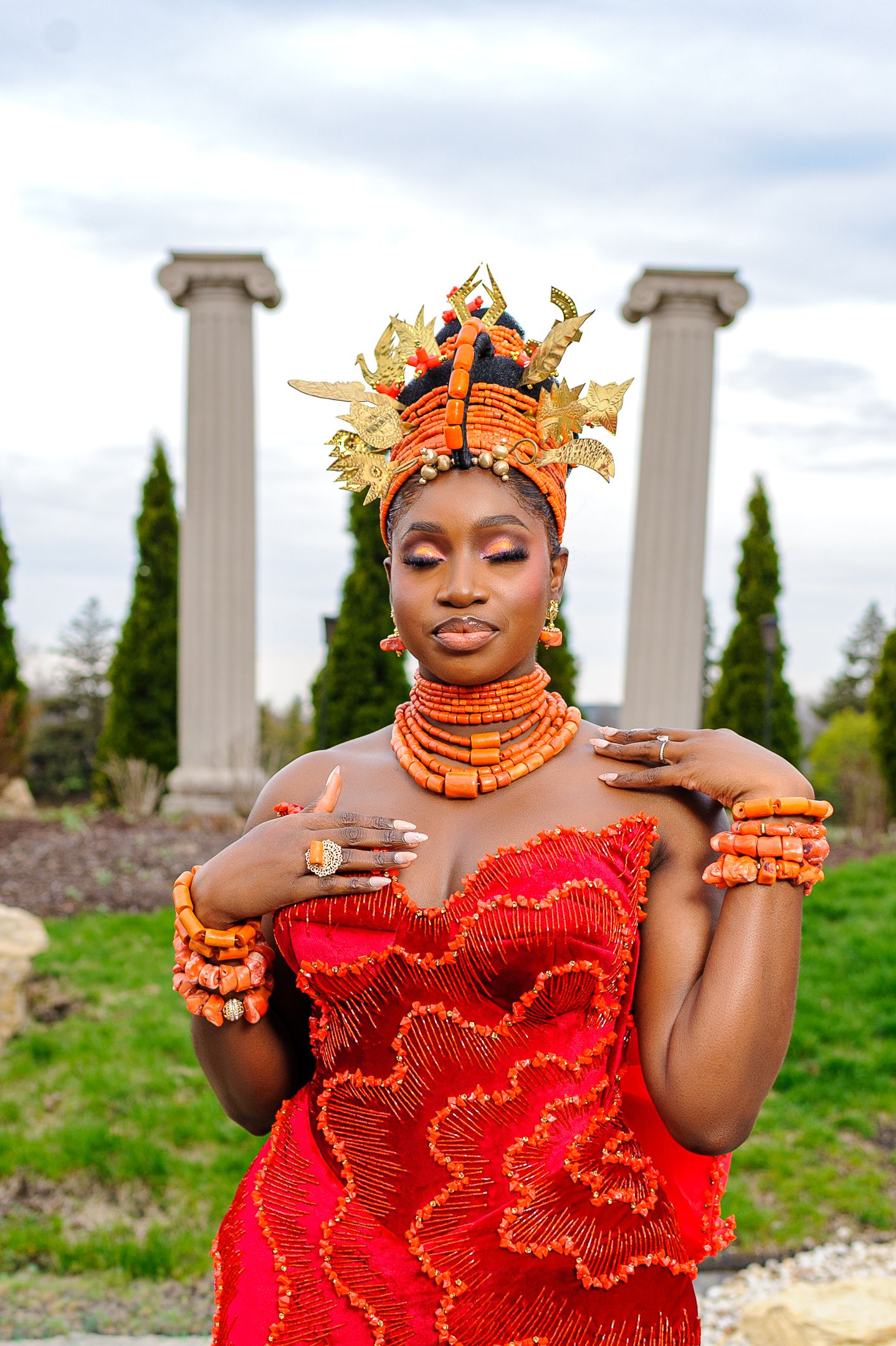 Nigerian bride in red beaded gown and coral crown poses gracefully before stone columns.