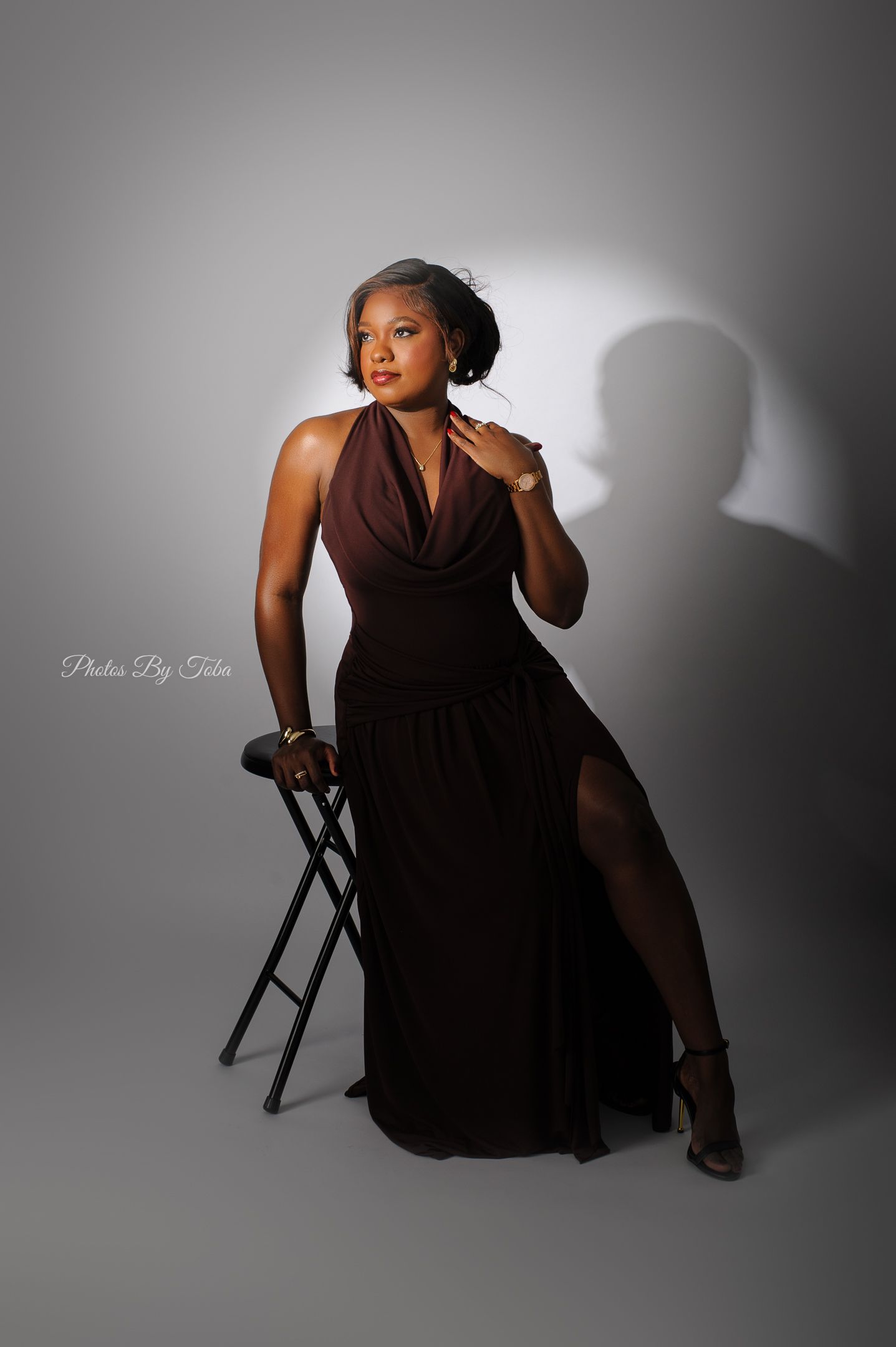 Woman in elegant brown halter gown seated on stool, posing gracefully in professional studio photography session.