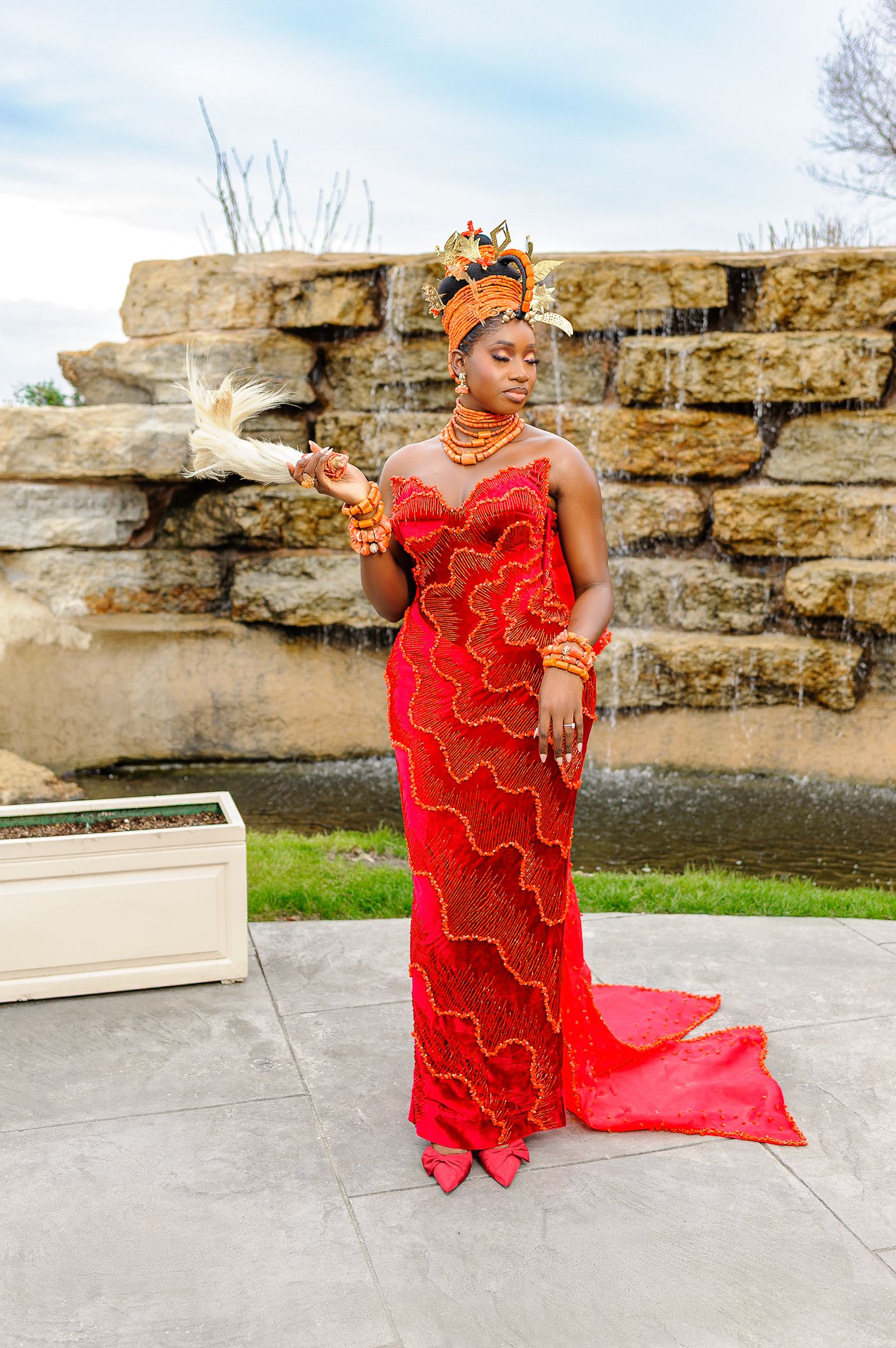 Woman in red beaded Nigerian traditional wedding gown with gold headdress holding a white dove outdoors.