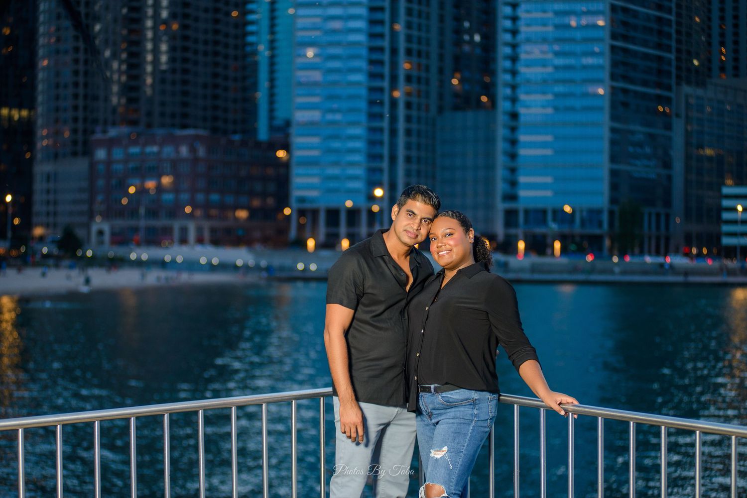 Couple posing together at night by a waterfront railing with illuminated city skyscrapers in the background.