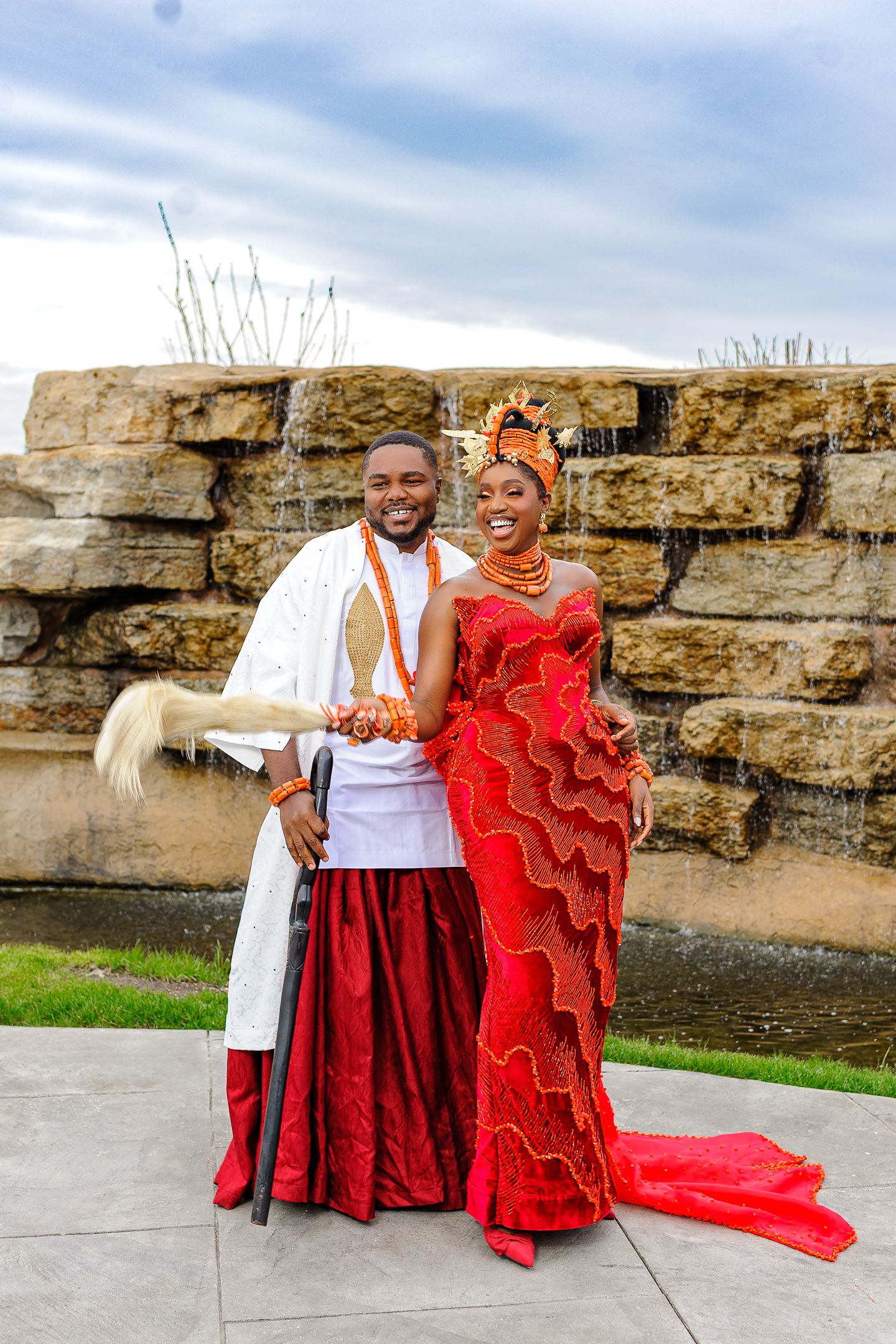 Couple in matching red and white Nigerian traditional wedding attire posing together outdoors near stone wall.