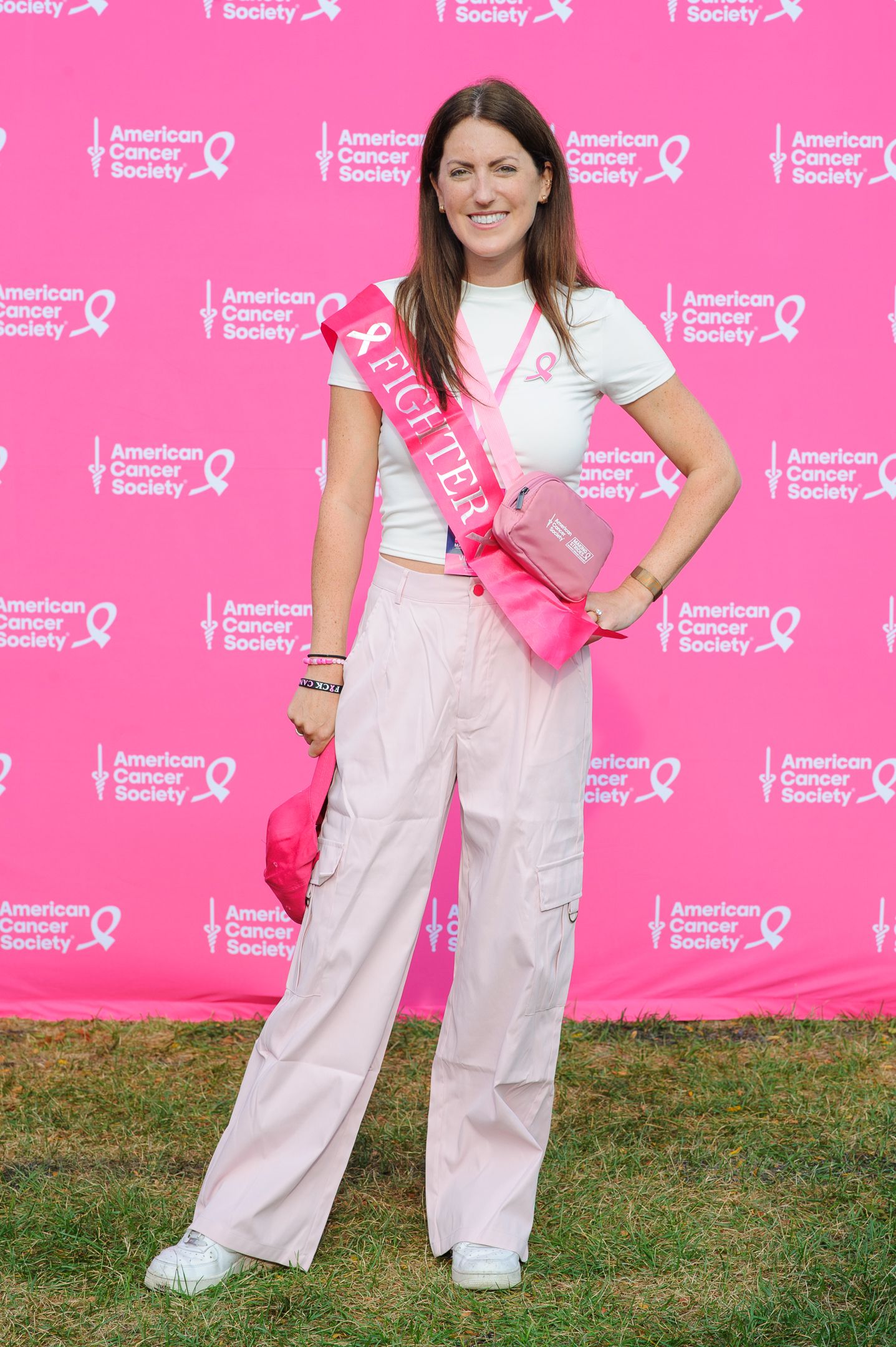 Person in white pants and t-shirt poses against pink backdrop with breast cancer awareness ribbons and logos.
