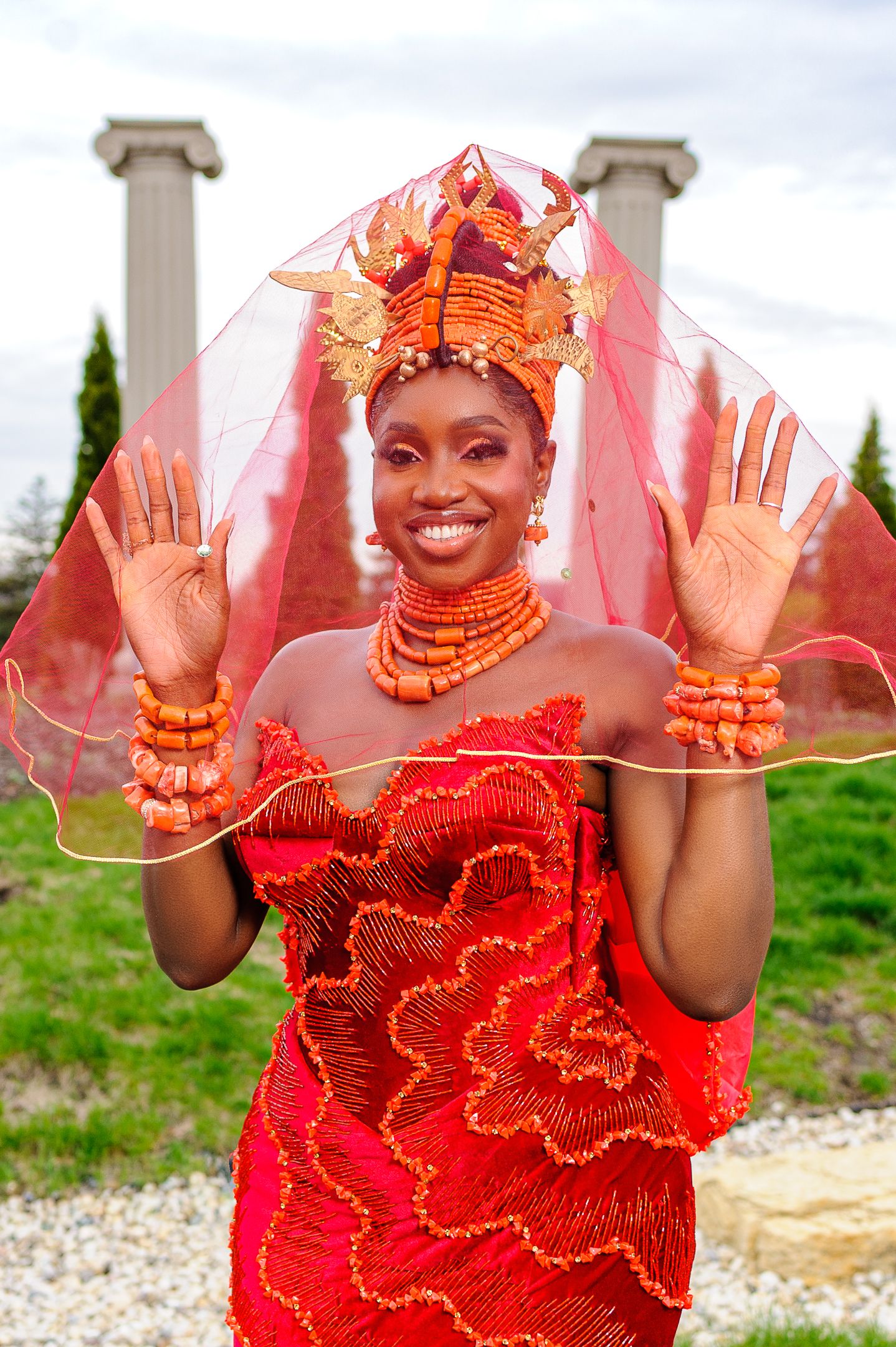 Nigerian bride in red traditional dress and coral crown lifts sheer veil with both hands.