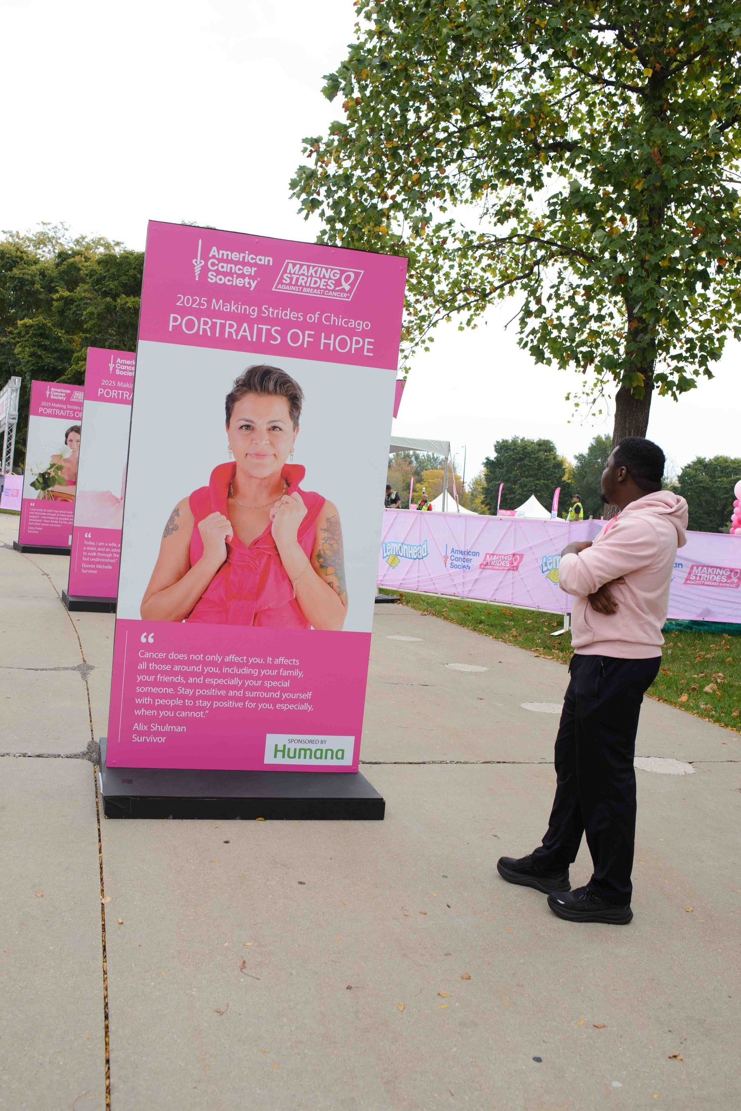 Pink promotional banner for breast cancer awareness stands on sidewalk near trees while person views display.
