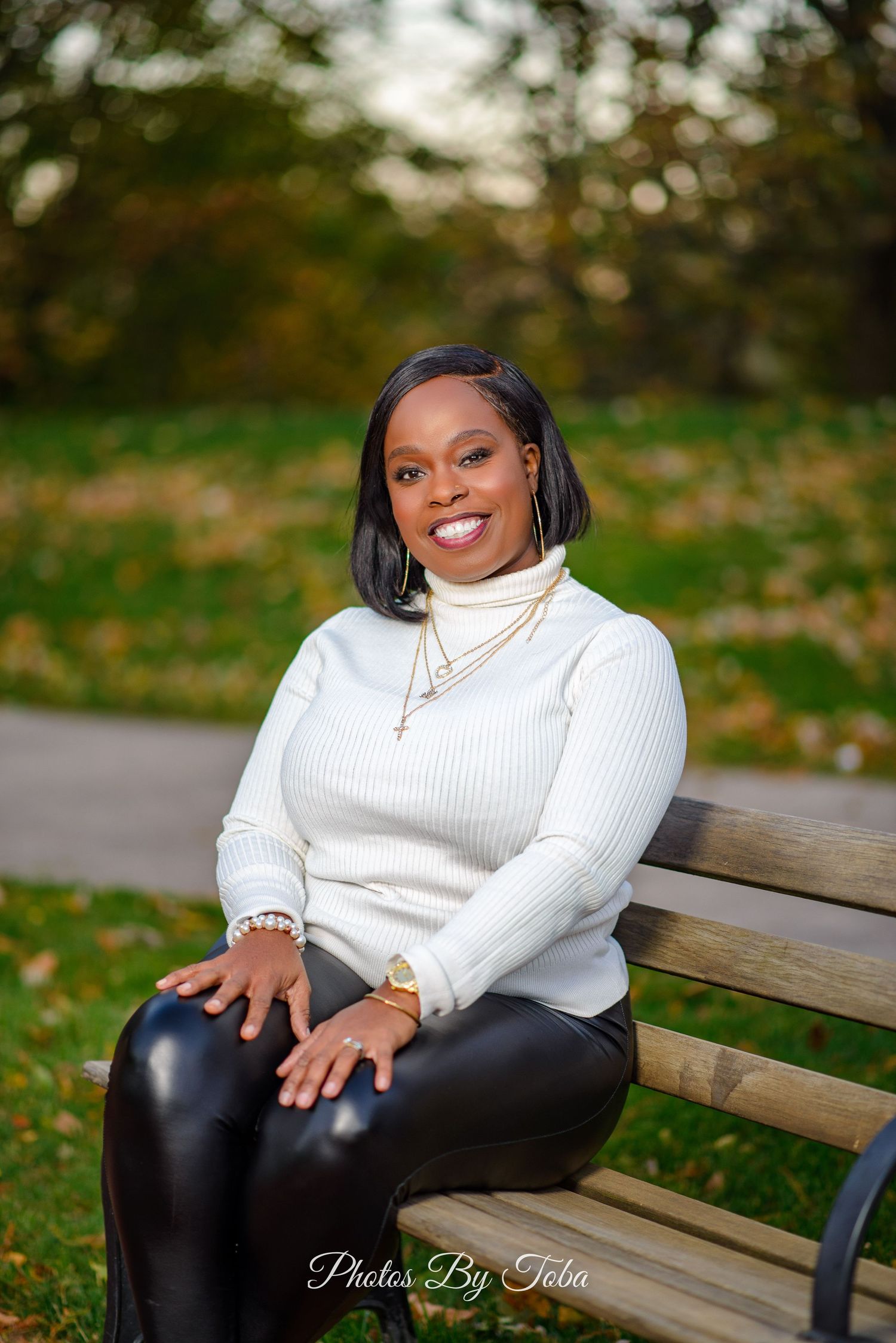 A person in a white sweater and black pants sits on a wooden bench outdoors during autumn.