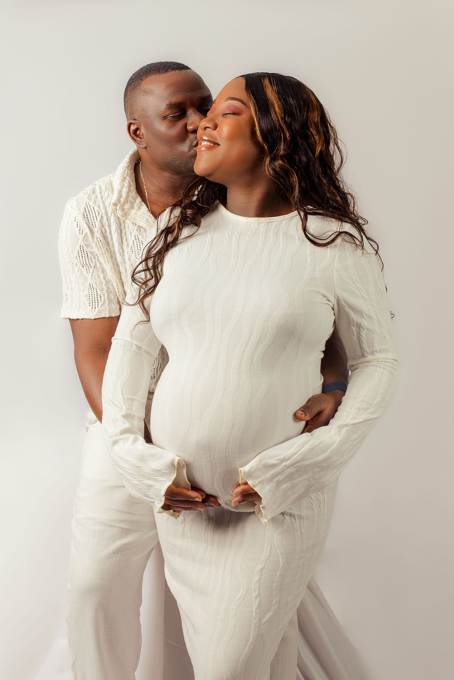 Maternity photoshoot with couple wearing matching white outfits against light background.
