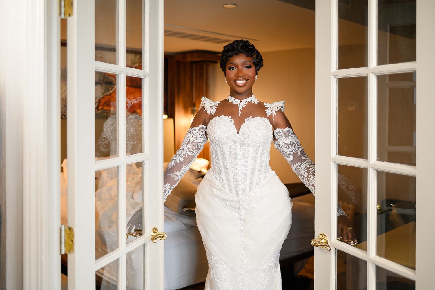 Bride in elegant lace mermaid wedding gown with long sleeves posing in French doorway, smiling radiantly.