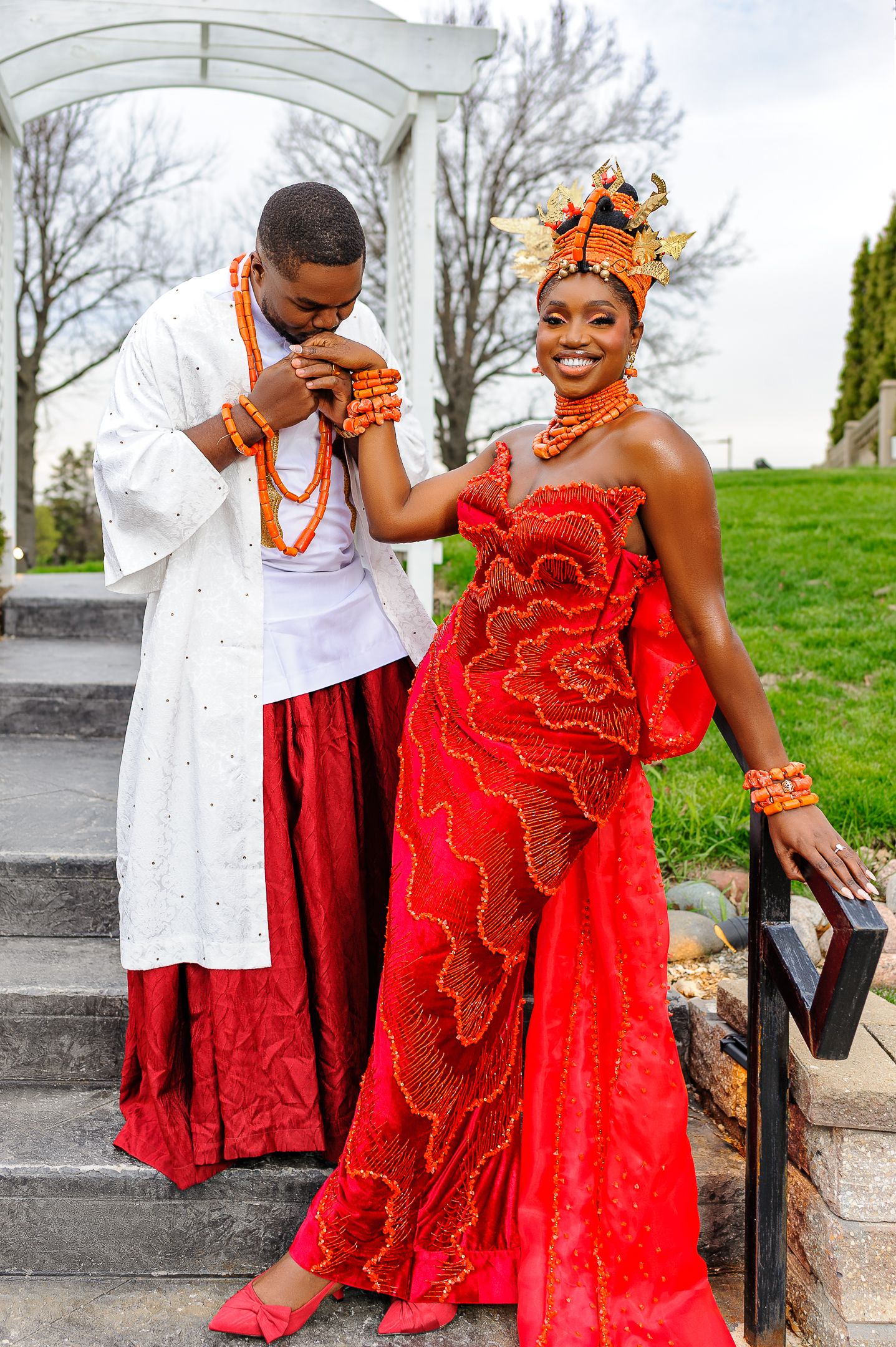 Couple in Nigerian traditional wedding attire, man in white agbada kisses womans hand in red lace gown and coral beads.