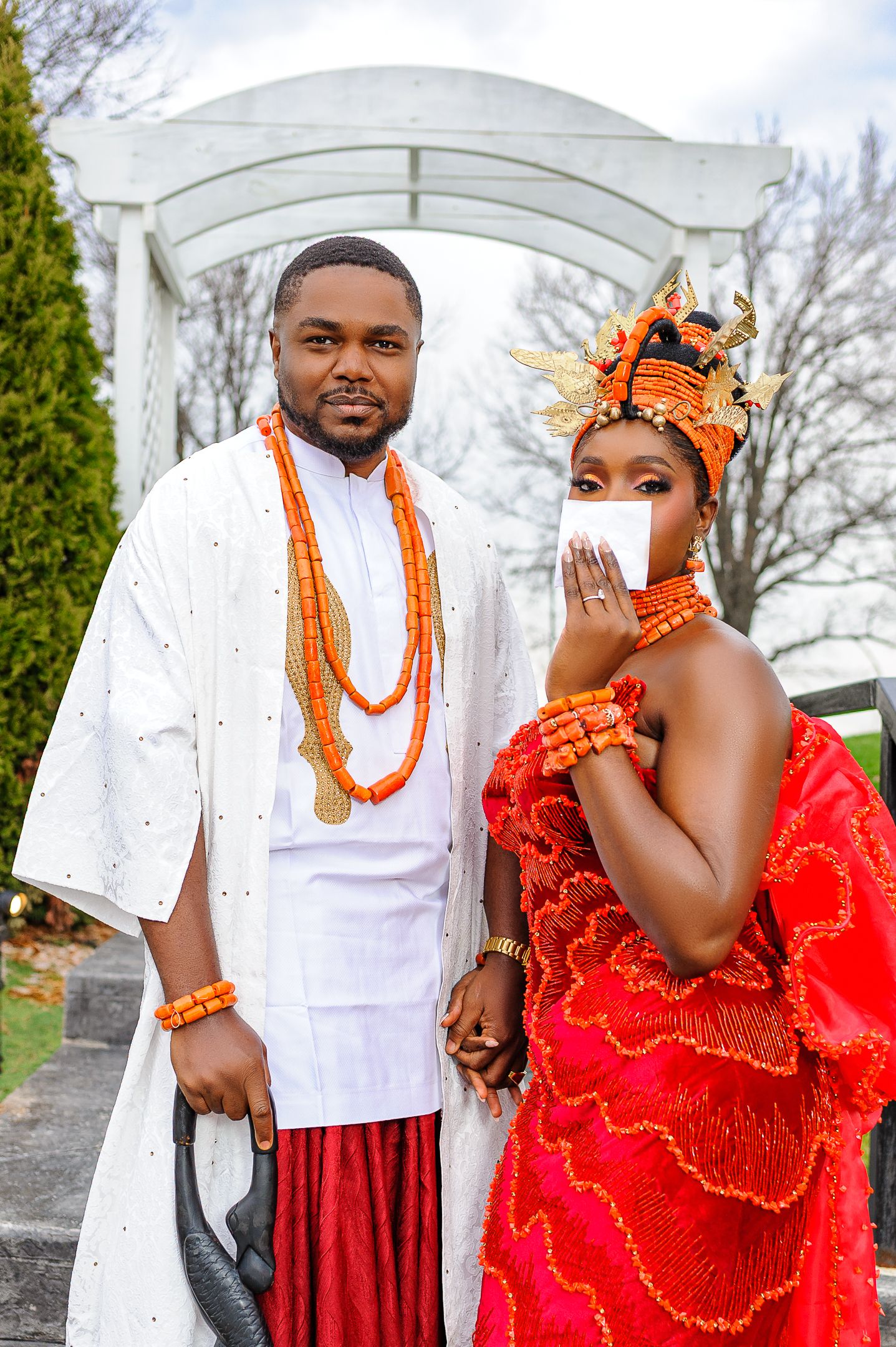 Nigerian couple in traditional Urhobo wedding attire, man in white agbada and woman in red coral beaded dress.