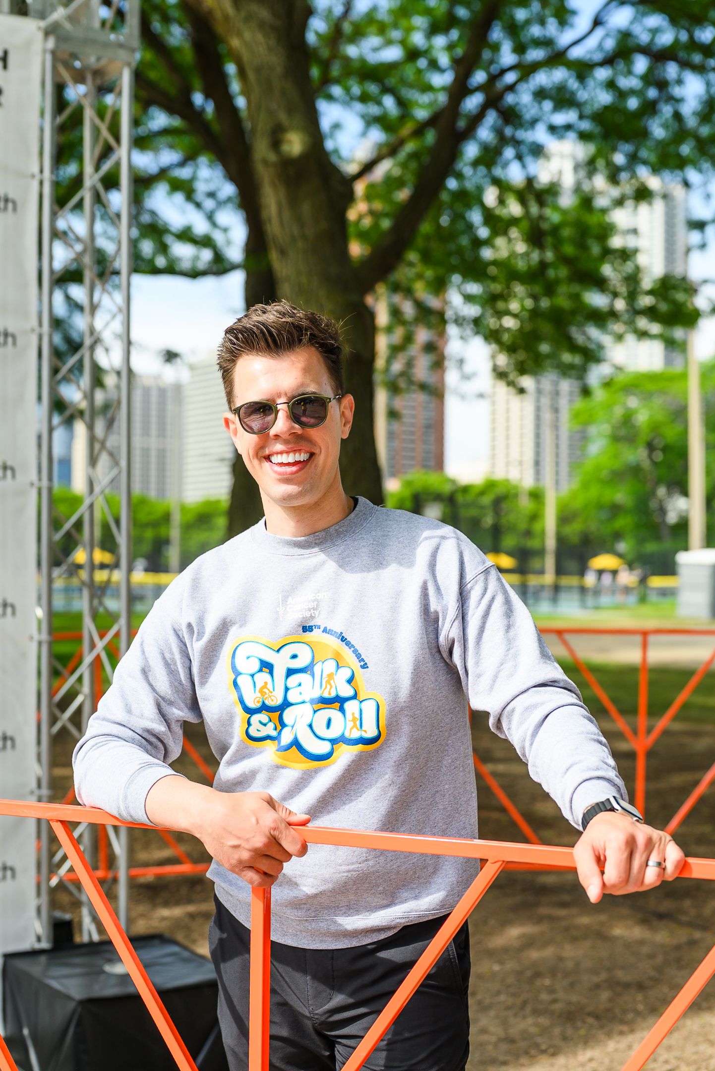Young man in Walk and Roll sweatshirt smiles leaning on orange barrier at outdoor charity event.