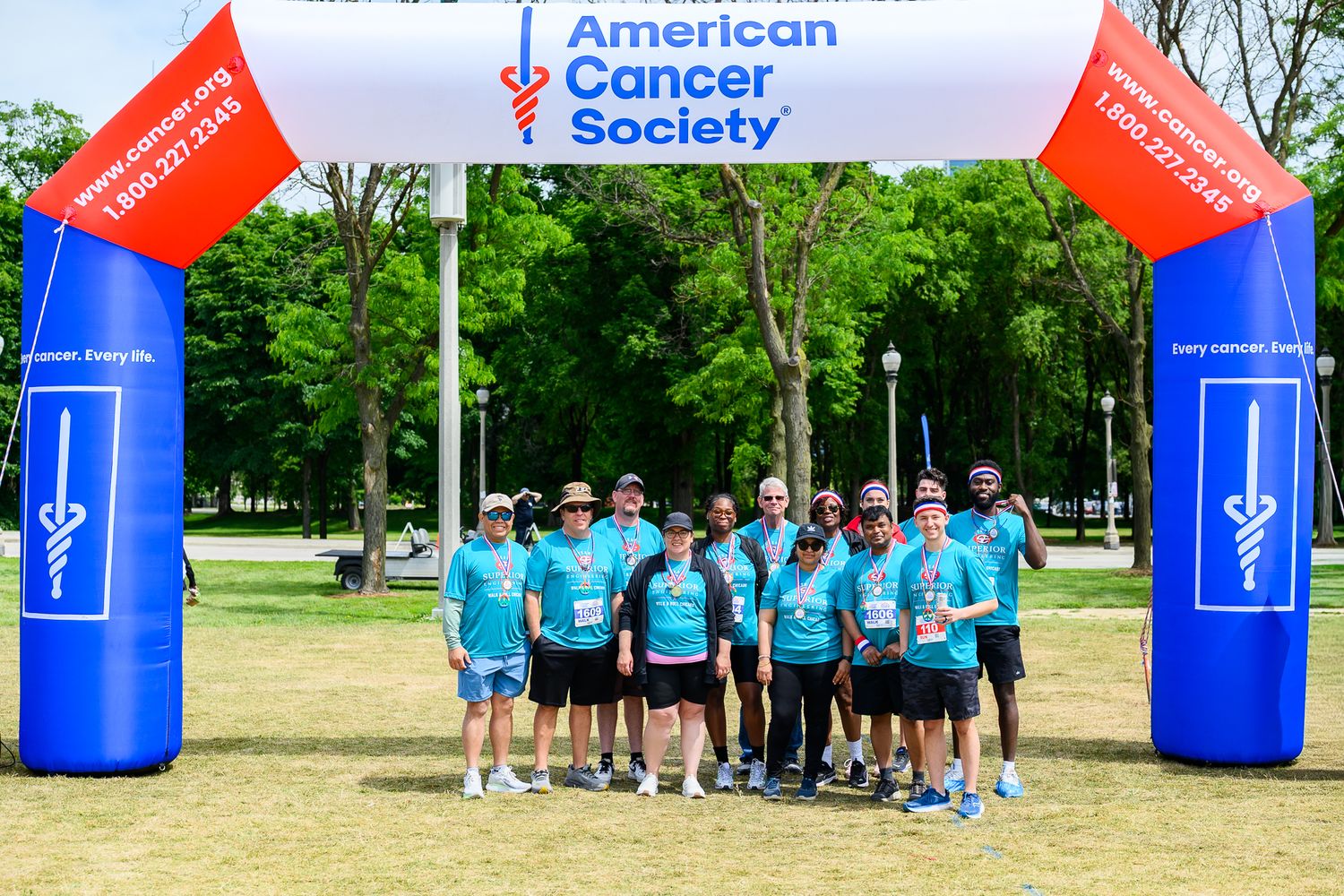 Group of people in teal shirts posing under American Cancer Society arch at outdoor charity walk event.
