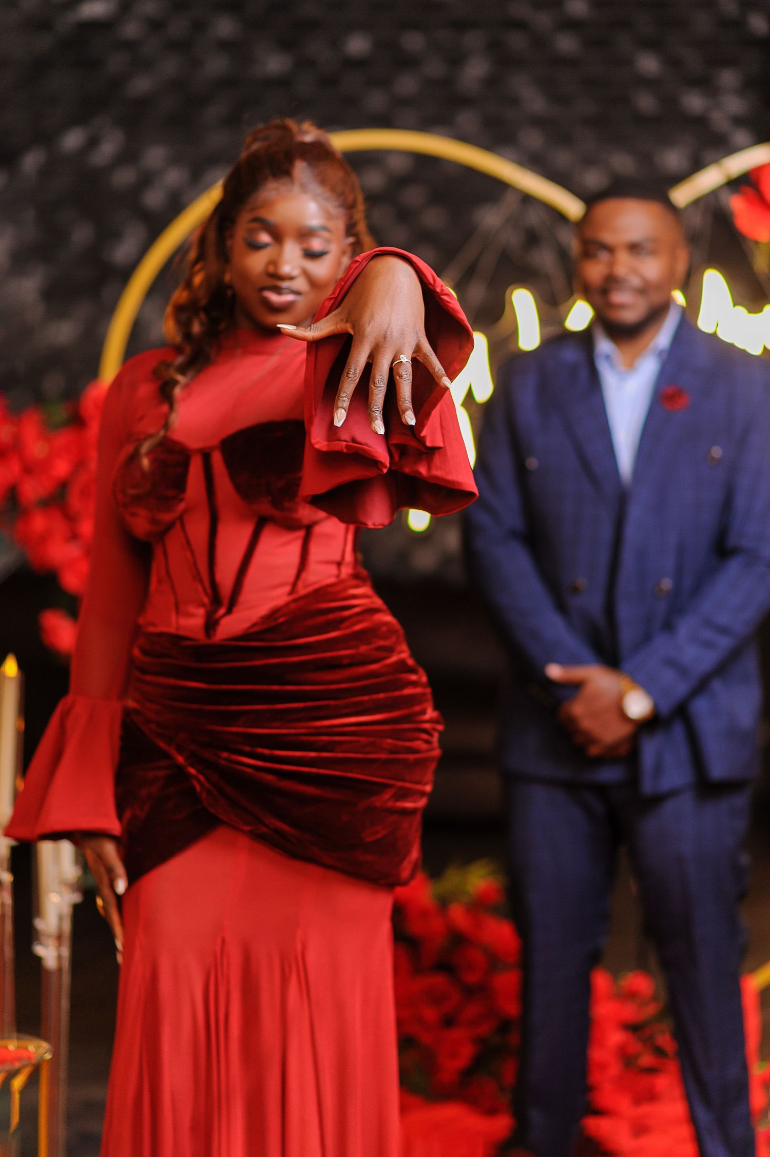 Woman in elegant red velvet ruffle gown posing at romantic proposal setup with man in blue suit smiling behind her.