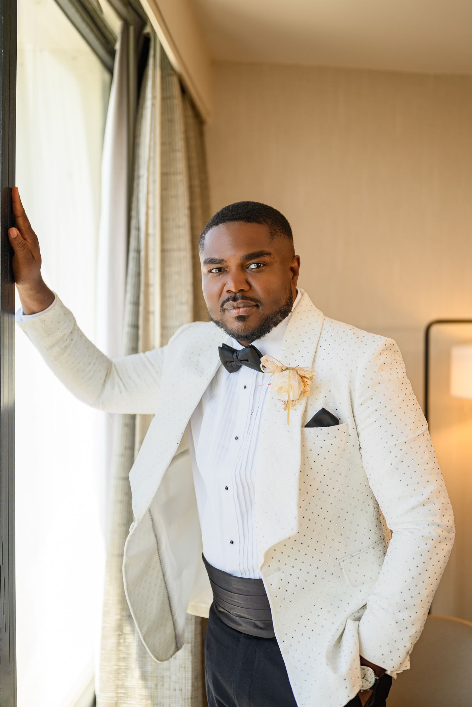Groom in white tuxedo jacket with bow tie and floral boutonniere poses by window on wedding day.