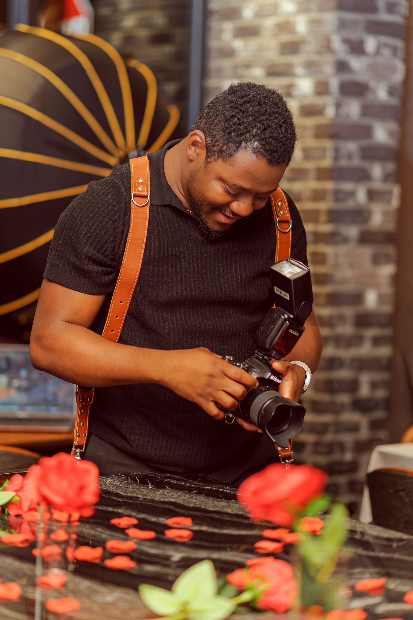 A photographer in a black shirt reviews shots on his camera, wearing a leather strap, surrounded by red roses.
