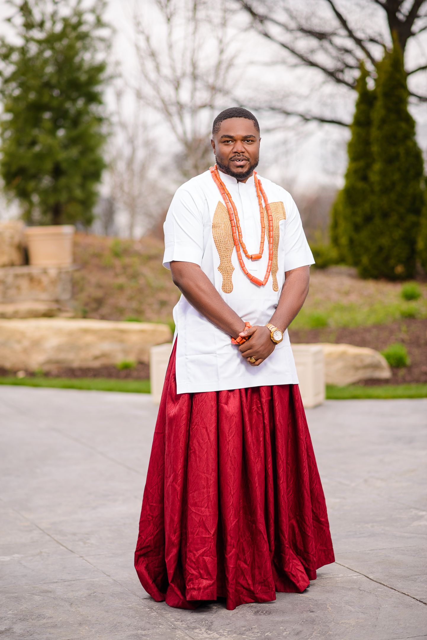 Nigerian groom in traditional white embroidered top and red wrapper with coral bead necklace at outdoor wedding.