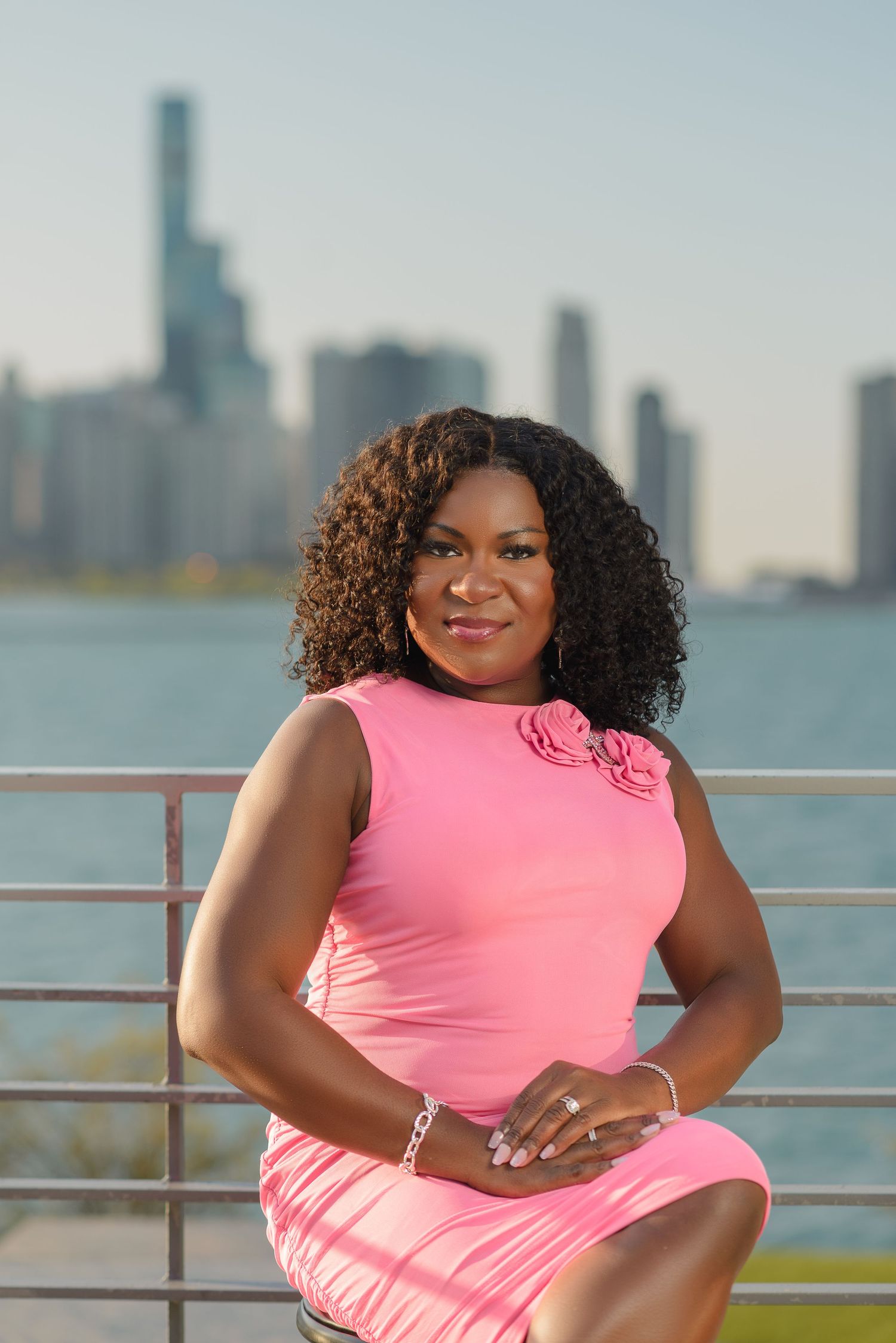 A person in a pink dress poses against a cityscape backdrop near a waterfront railing.