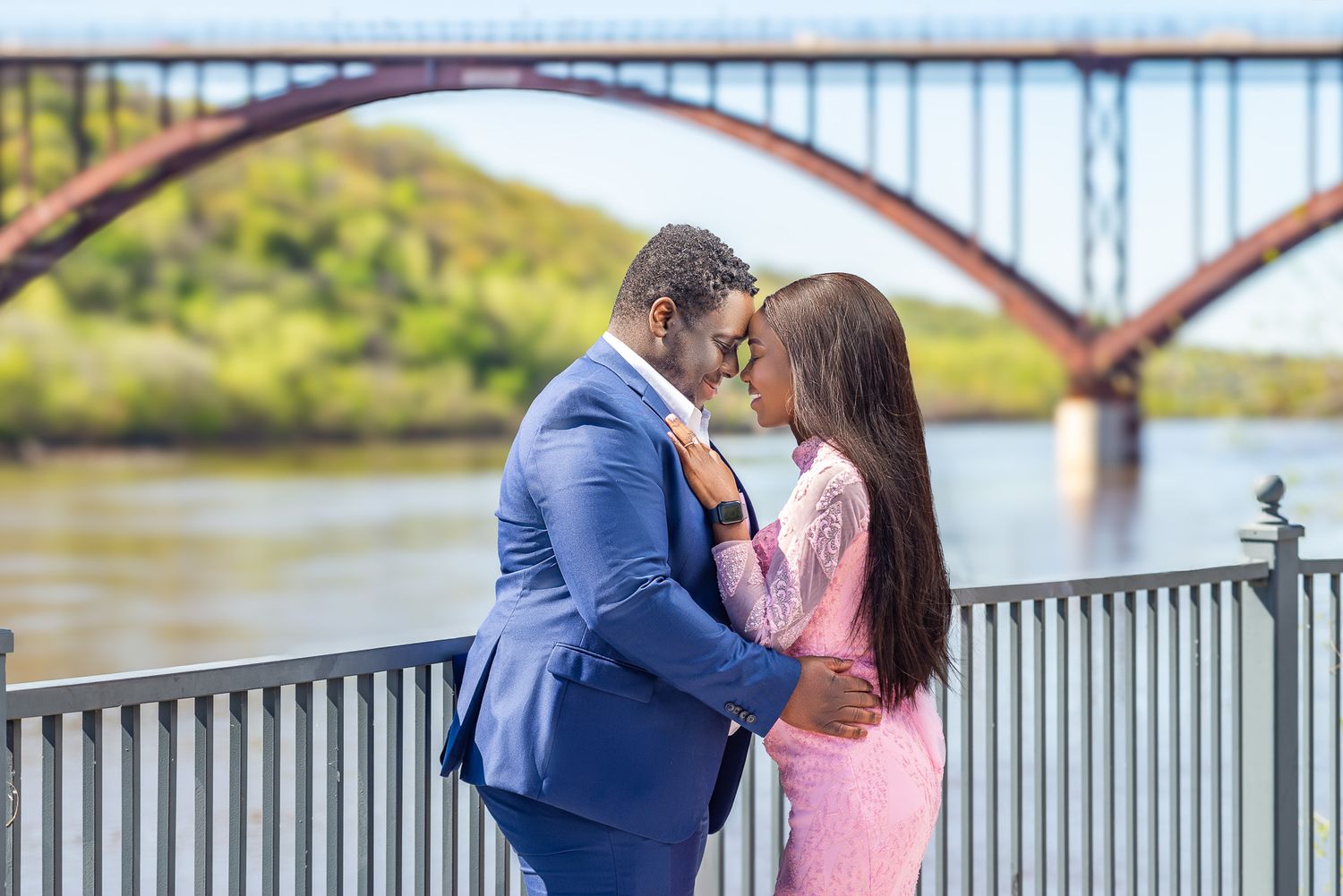 Couple in formal wear sharing a romantic moment by a river with a red arch bridge in the background.