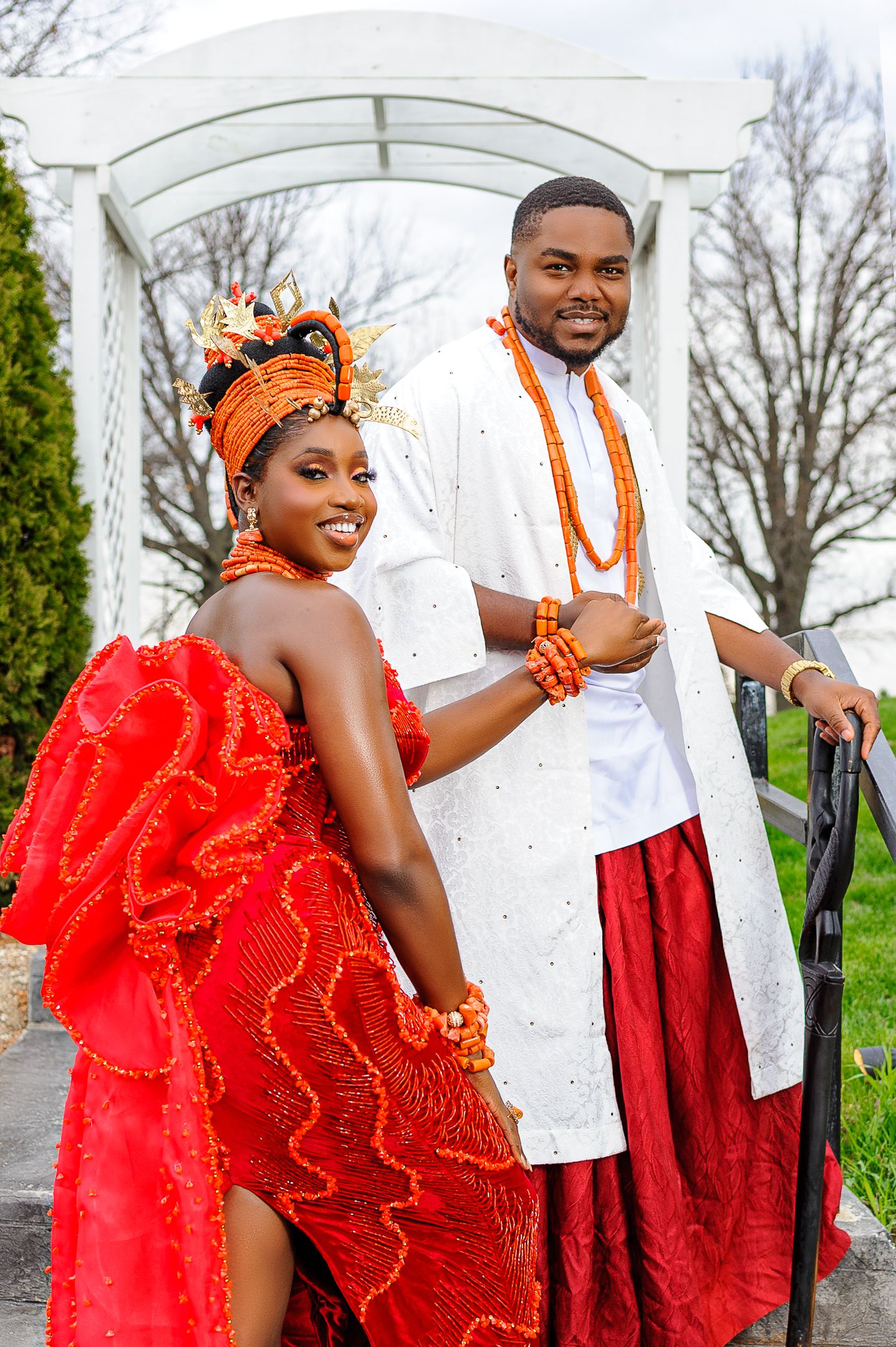 Couple in vibrant Nigerian traditional wedding attire with coral beads and red garments pose under a white gazebo.