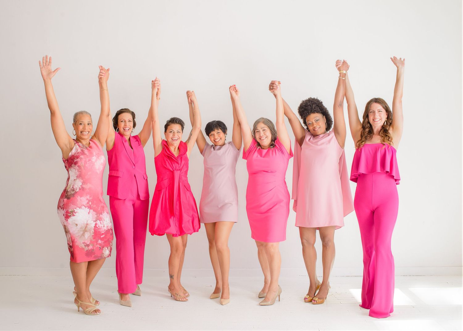A group of people wearing pink outfits raise their arms joyfully in celebration against a white studio backdrop.