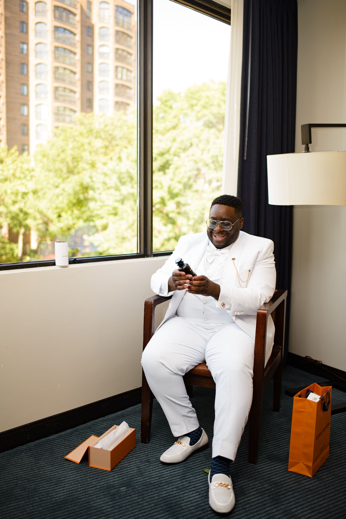 Person in all-white outfit sitting casually in hotel room with large windows.