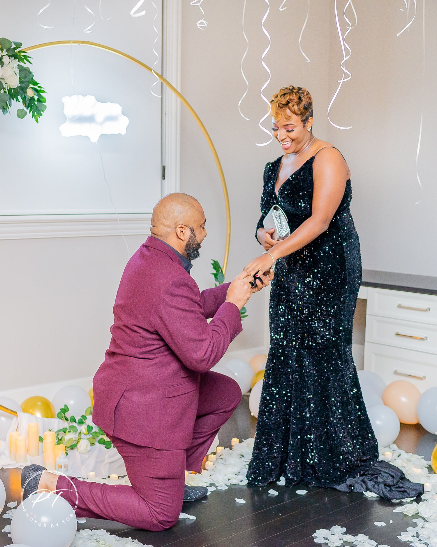 Man in burgundy suit proposes to woman in black sequin gown surrounded by candles, flowers, and balloons.