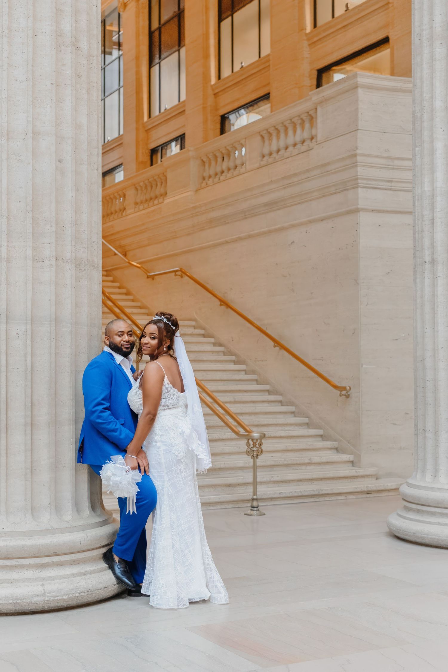 Bride in white lace gown and groom in blue suit pose elegantly by marble columns and staircase in grand building.