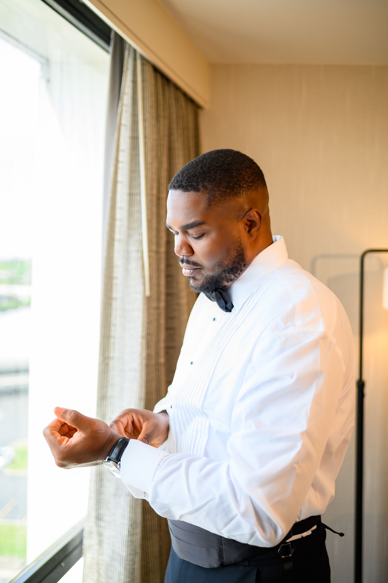 A well-dressed Black groom adjusts his cufflinks by a bright hotel window, wearing a white shirt and black bow tie.