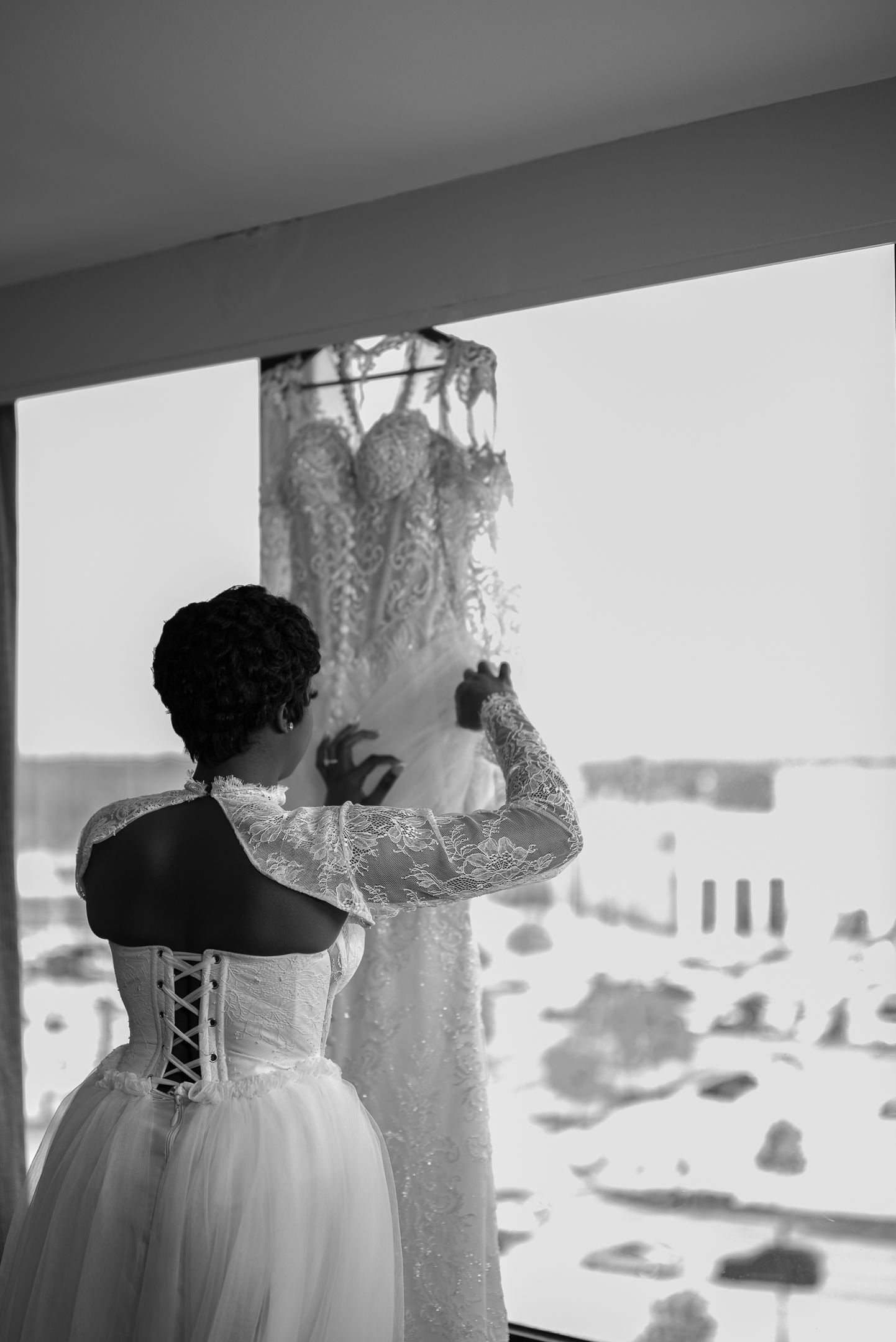 Black and white photo of a bride in a lace corset gown admiring her wedding dress hanging by a window.