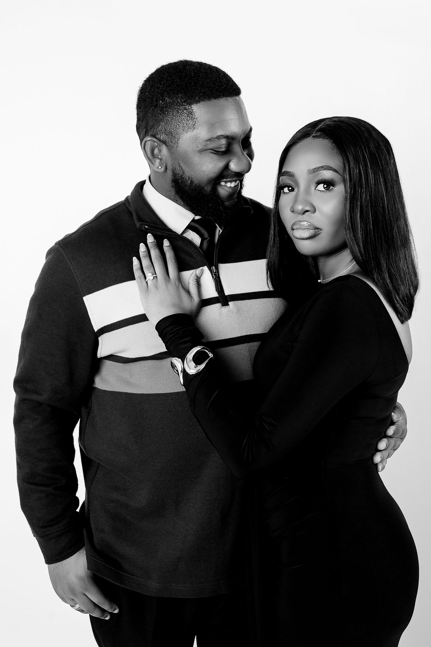Black and white portrait of a smiling couple embracing, man in striped sweater, woman in black dress, studio background.