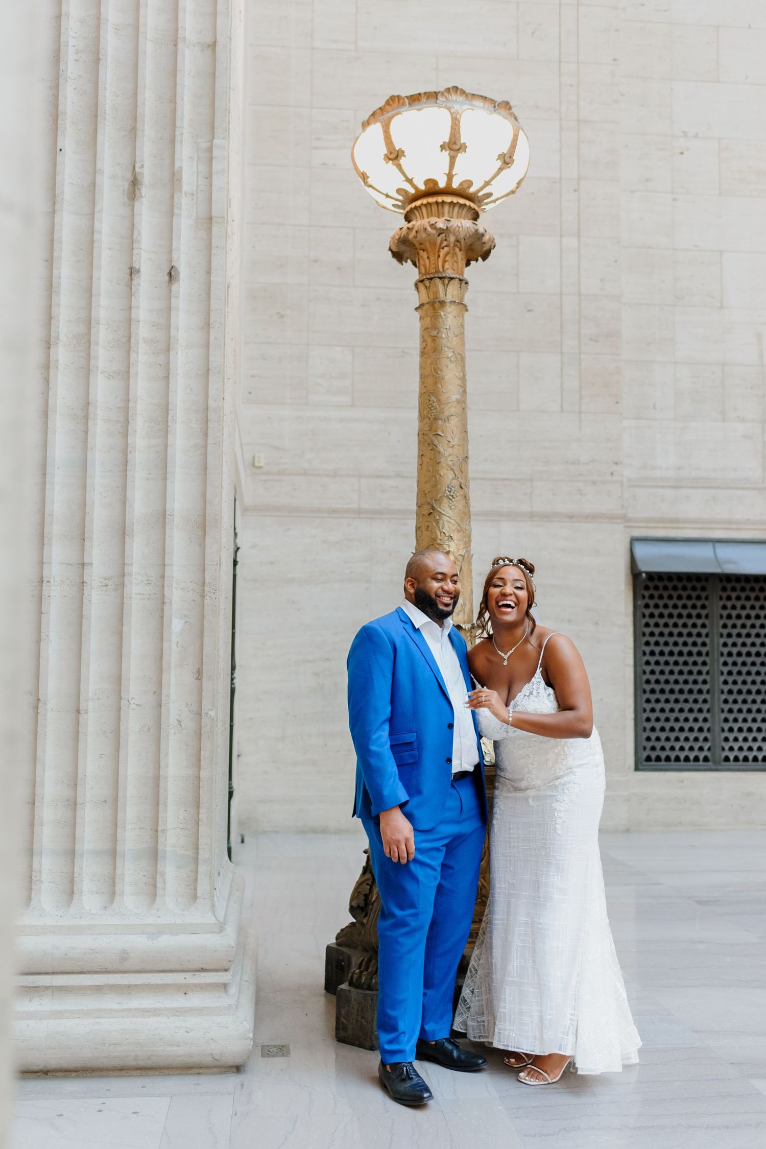 A couple in wedding attire poses against white stone columns at a grand architectural venue.
