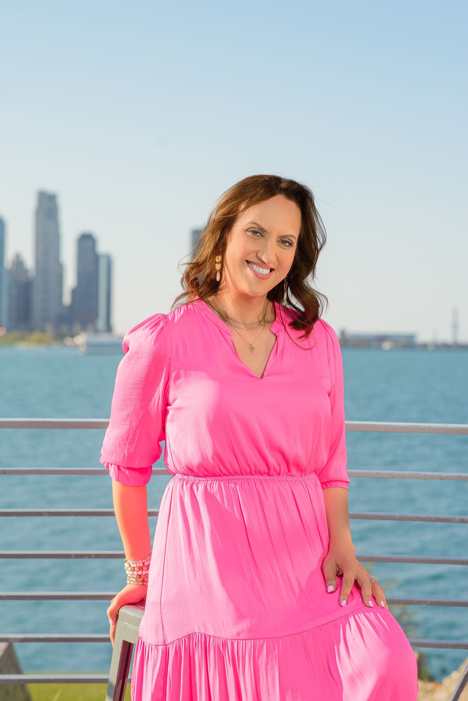 A woman in a bright pink tiered dress poses by the water with a city skyline in the background.