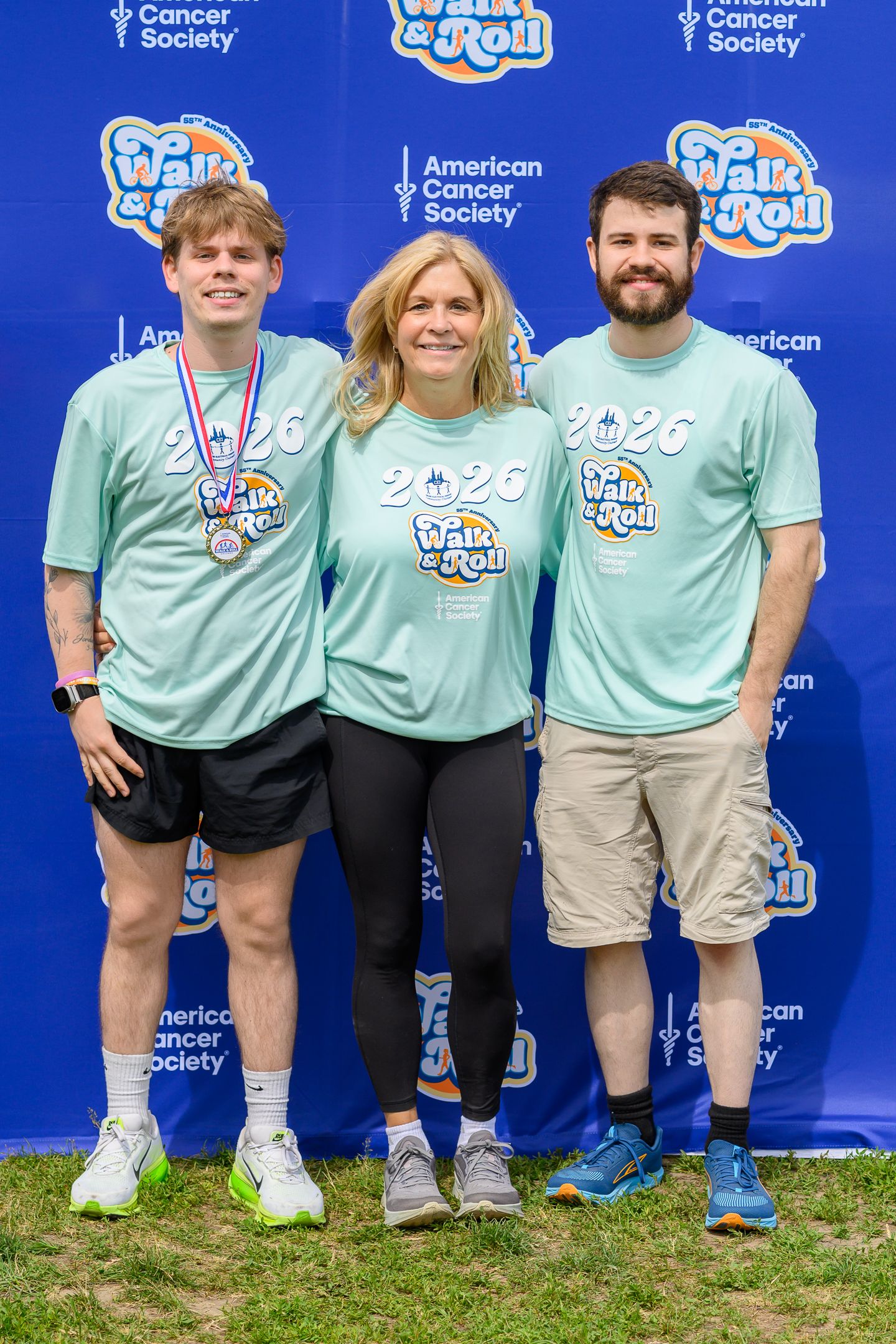 Three people in matching teal American Cancer Society Walk & Roll 2026 shirts pose together outdoors at charity event.