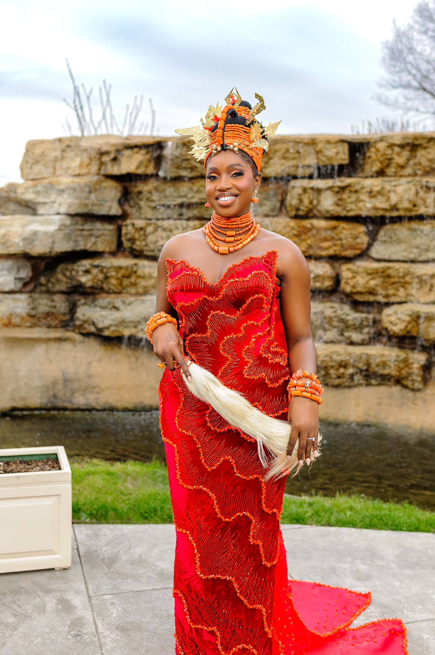 Nigerian bride in red beaded gown and ornate coral crown holds white feather fan at outdoor ceremony.
