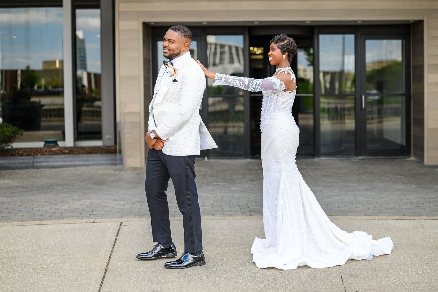 Bride in white gown adjusts grooms white tuxedo jacket outdoors before wedding ceremony.