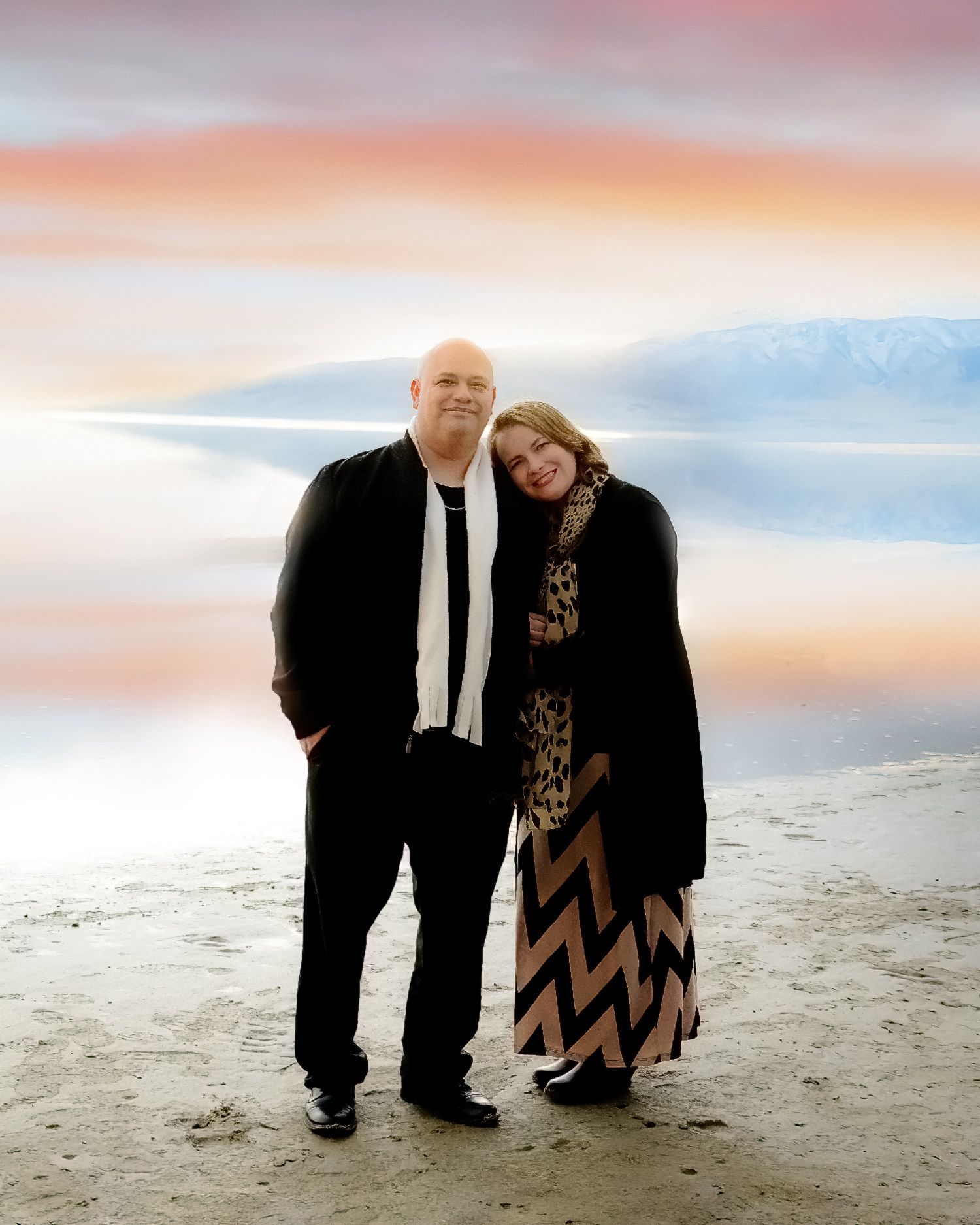 A couple standing close together on a beach during sunset with mountains in the distance.