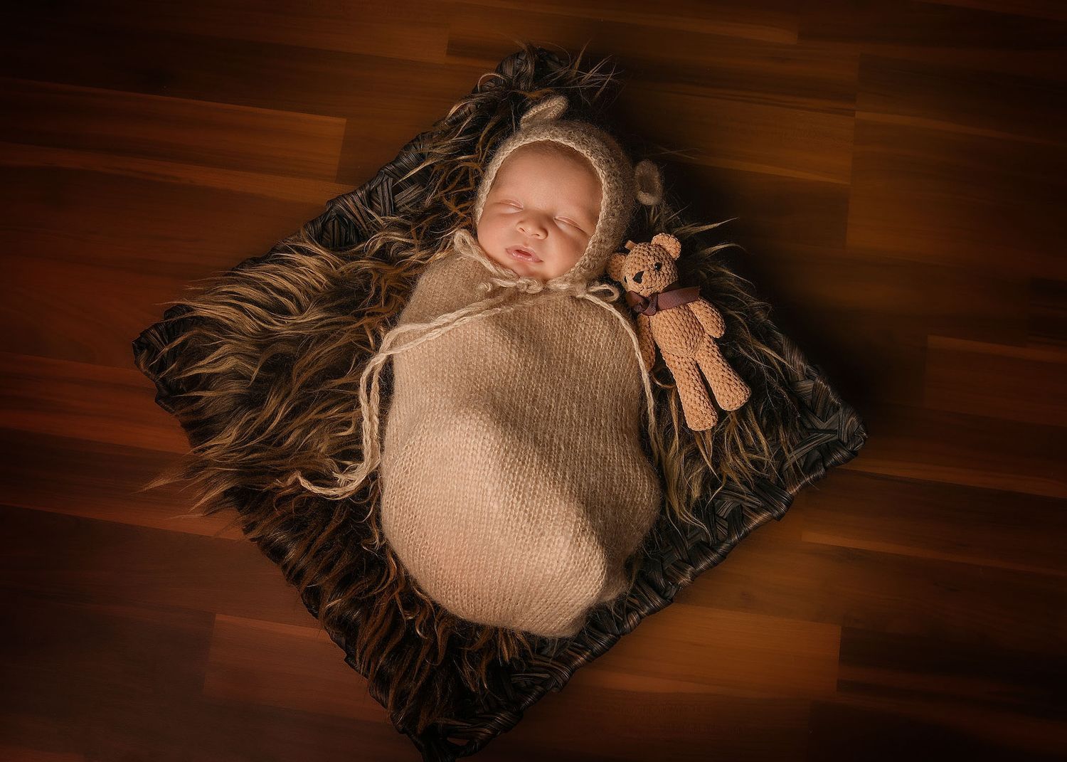 Newborn baby sleeping in brown knit wrap with teddy bear on dark fur rug, professional newborn photography.