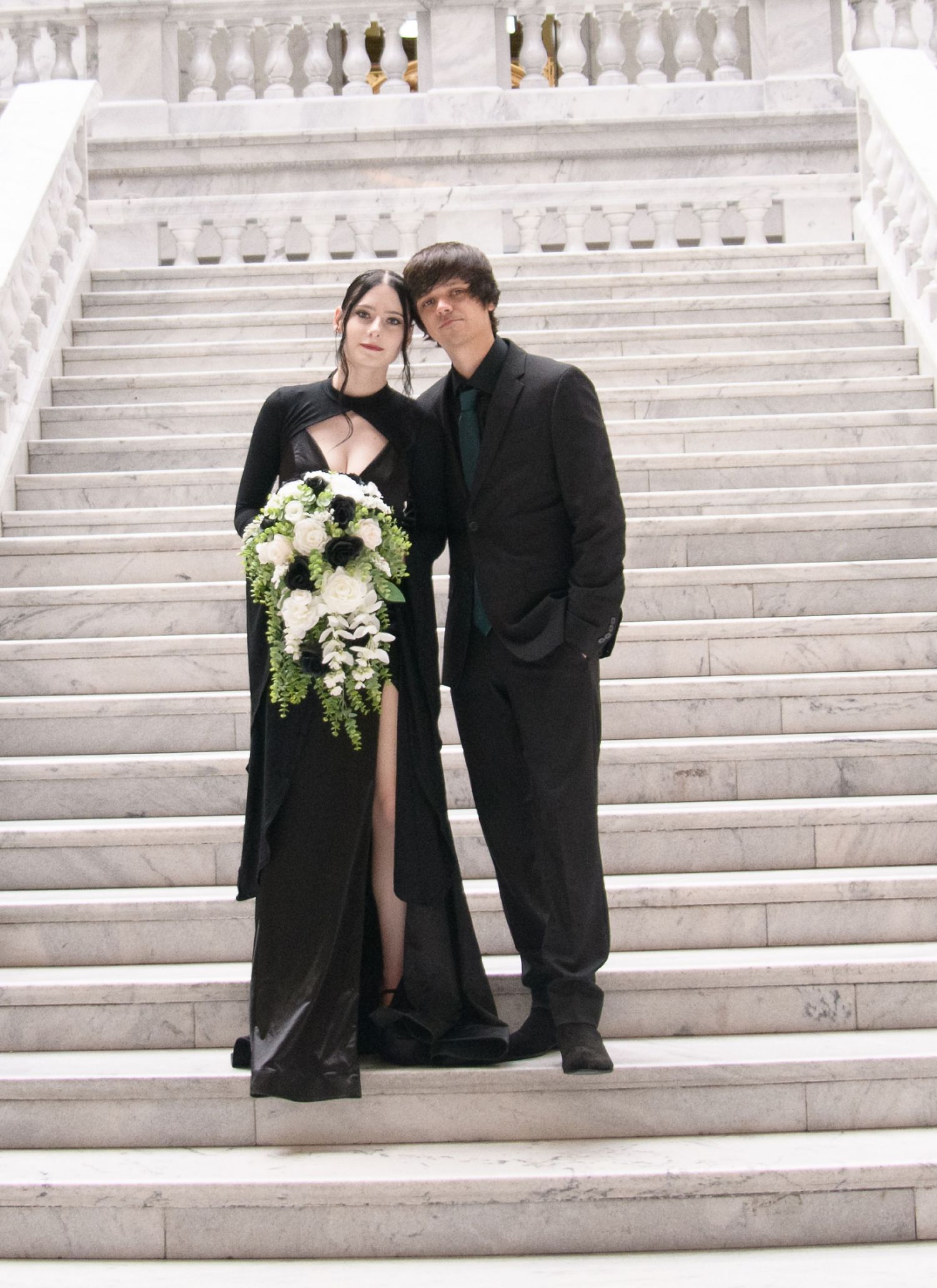 A couple in formal attire, with a black dress and suit, poses elegantly on white marble stairs.