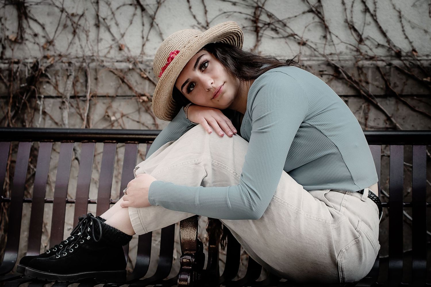 A moody portrait of someone in a sweater and hat sitting on a wooden bench against climbing vines.