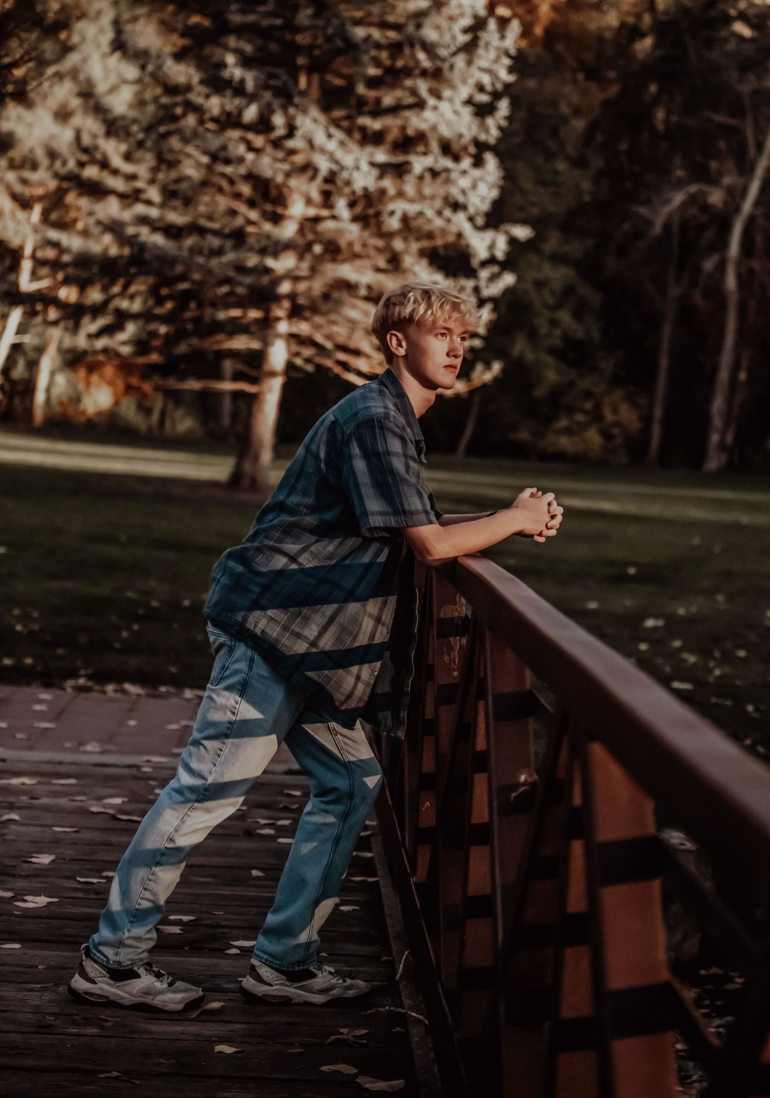 A youth leans against a wooden bridge railing in an autumn park setting.
