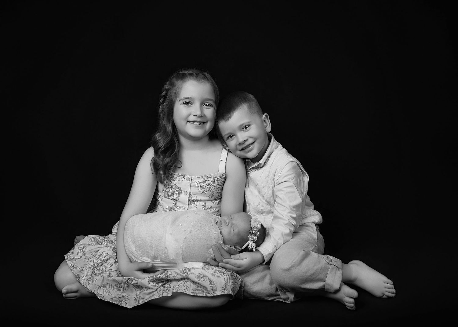 Black and white portrait of two young siblings, a girl in a dress and a boy in a shirt, sitting together smiling.