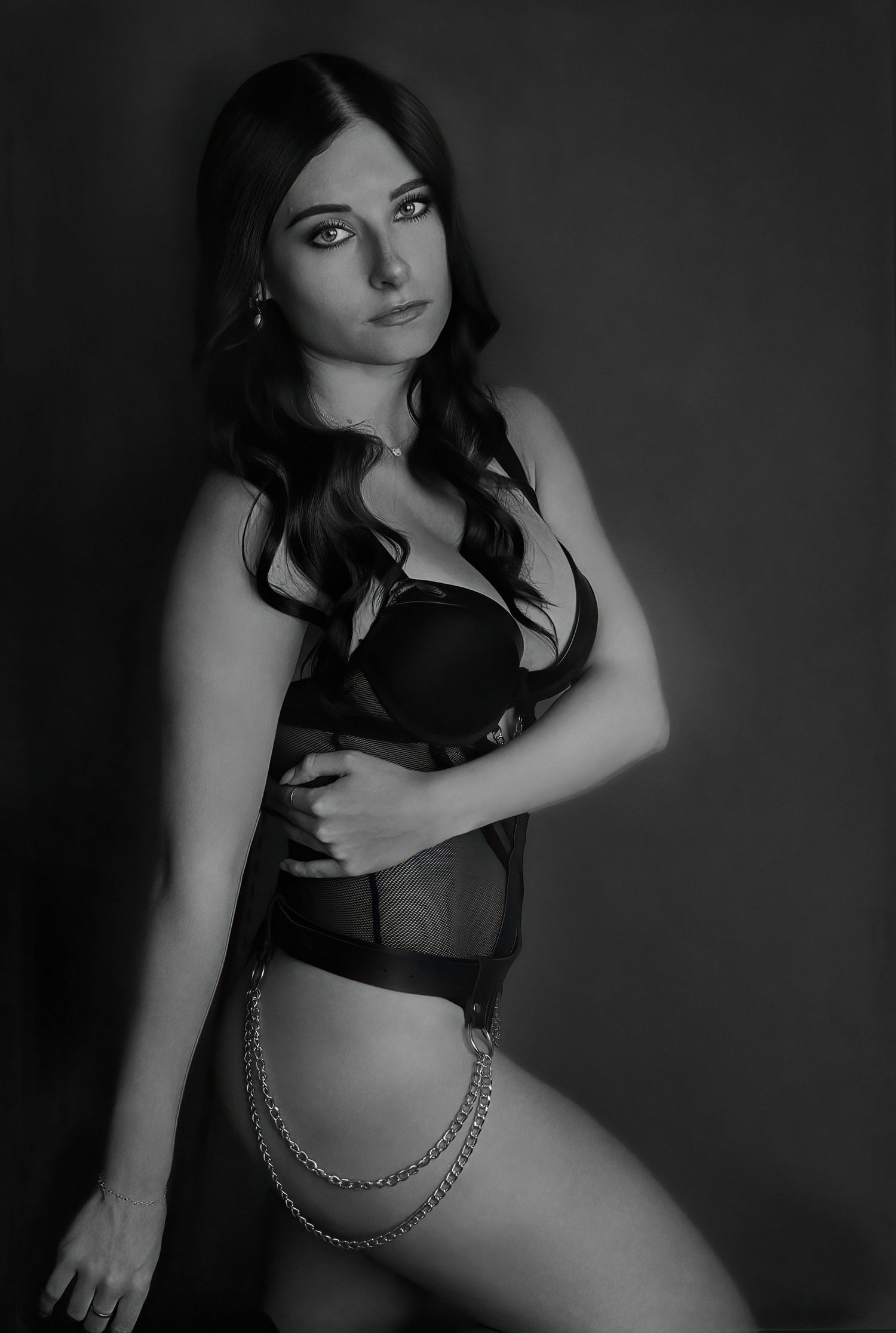 Black and white boudoir photo of a young brunette woman posing in black lingerie with chain accessory.