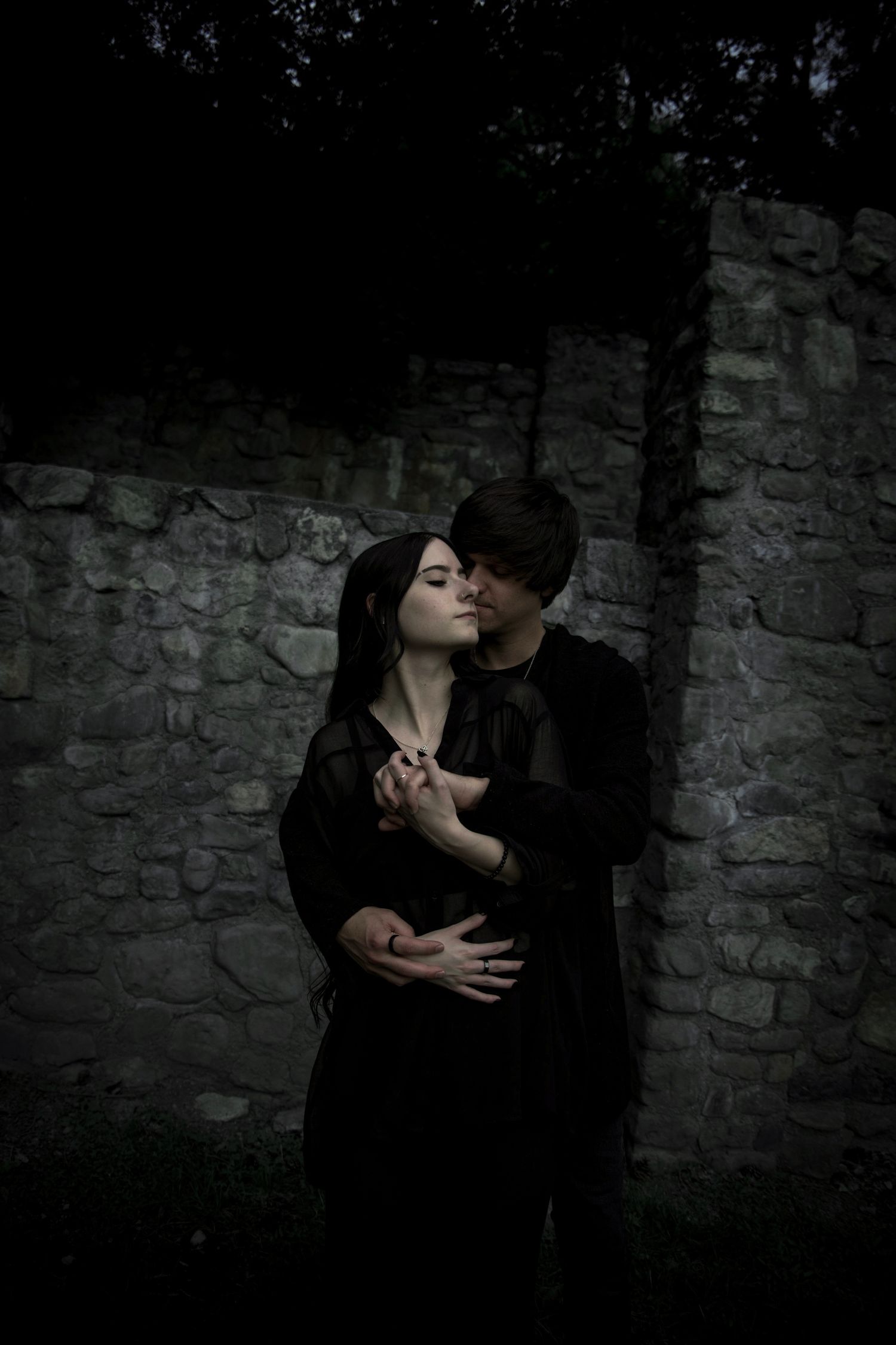 A romantic couple dressed in dark clothing embrace against a stone wall in a moody, atmospheric setting at dusk.