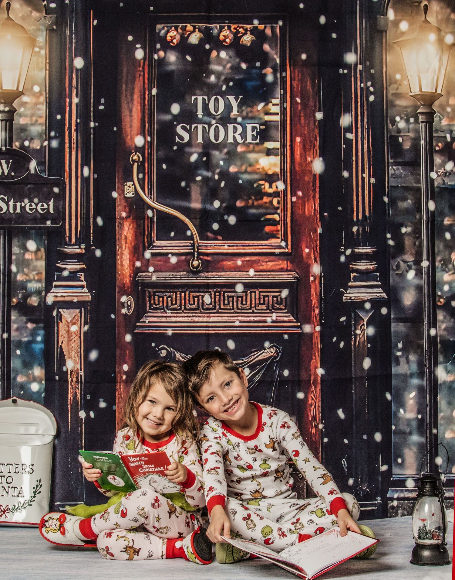 Children in matching holiday pajamas read a book together by a festive storefront window with falling snow.