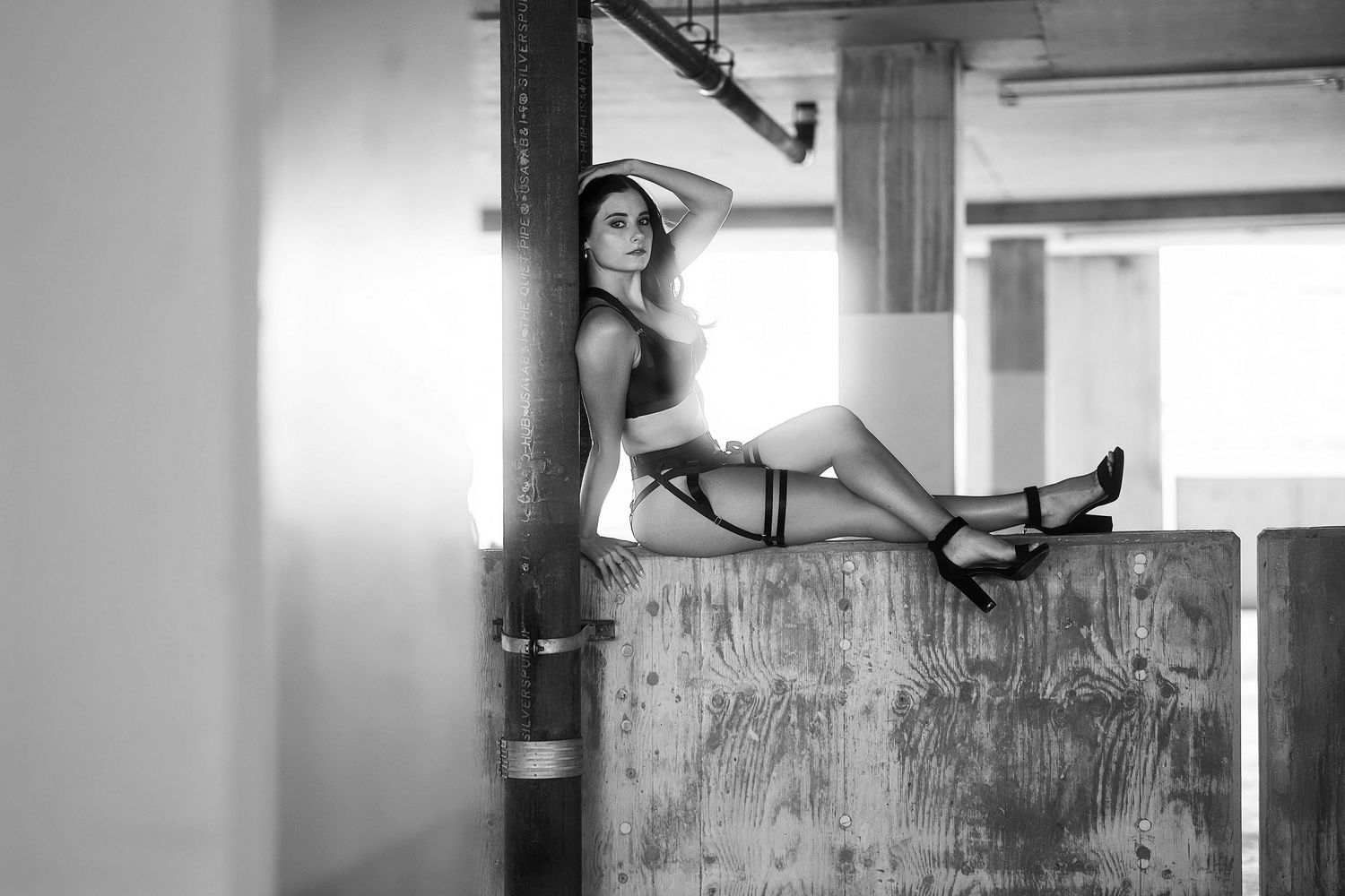 Black and white photo of a woman in lingerie posing on a concrete ledge in an industrial setting.