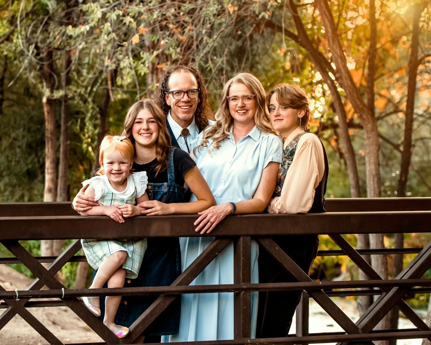 Family portrait on a wooden bridge during golden hour with sunlit trees in background.
