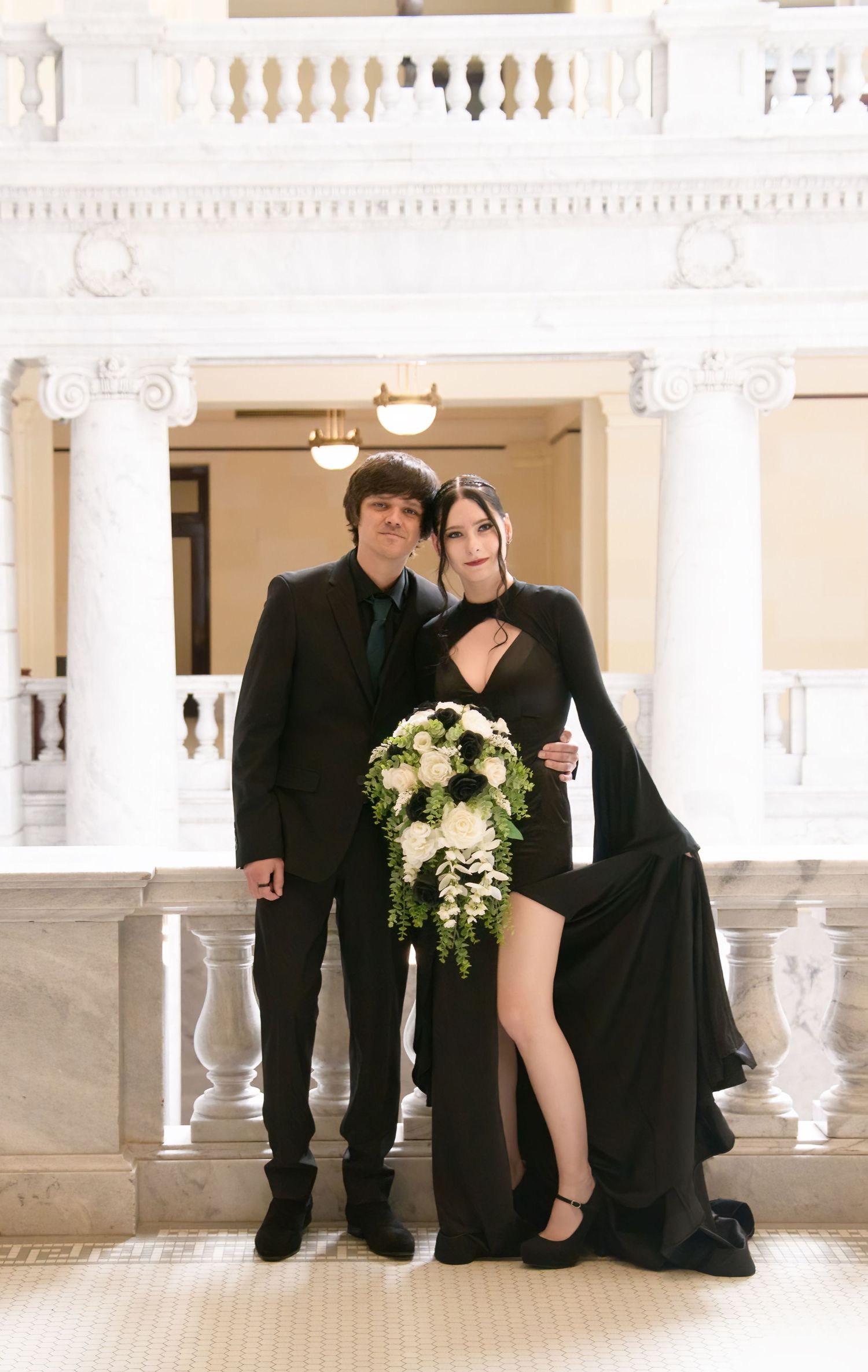 A wedding couple in all black formal attire poses together in a white marble corridor.