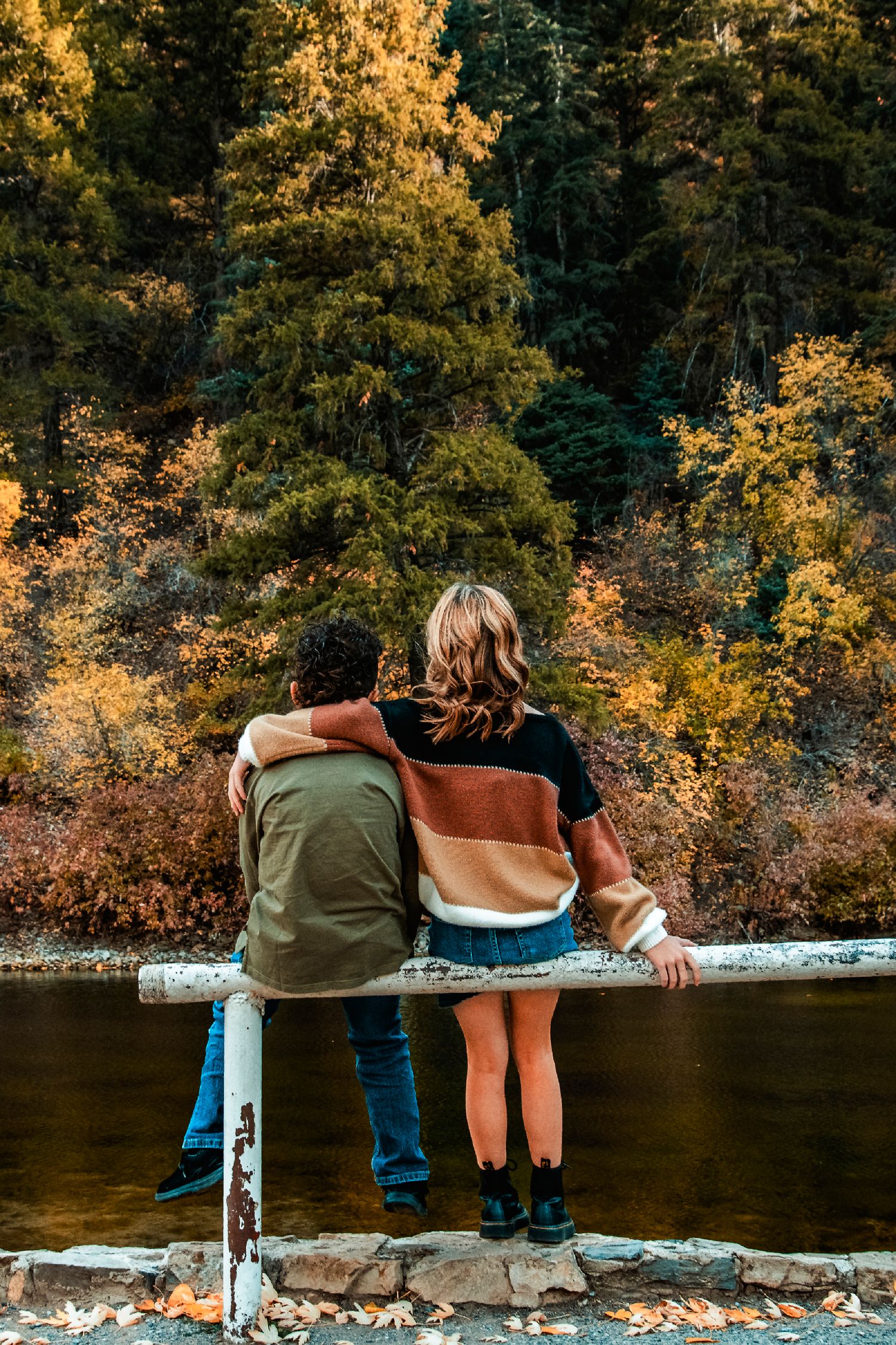 Two people sitting on railing overlooking a river surrounded by autumn trees in golden light.