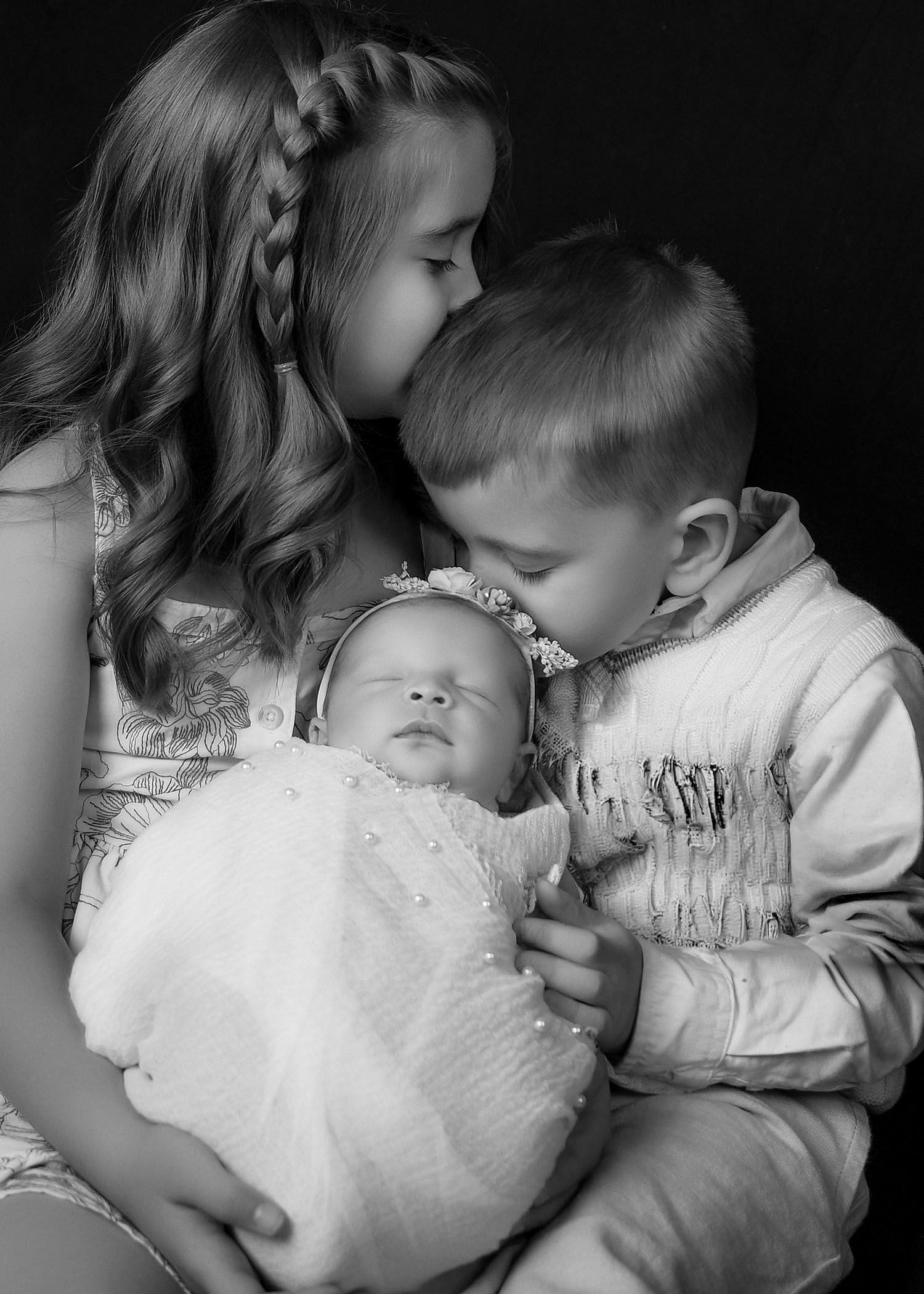 Black and white photo of two older siblings kissing a sleeping newborn baby wrapped in white.
