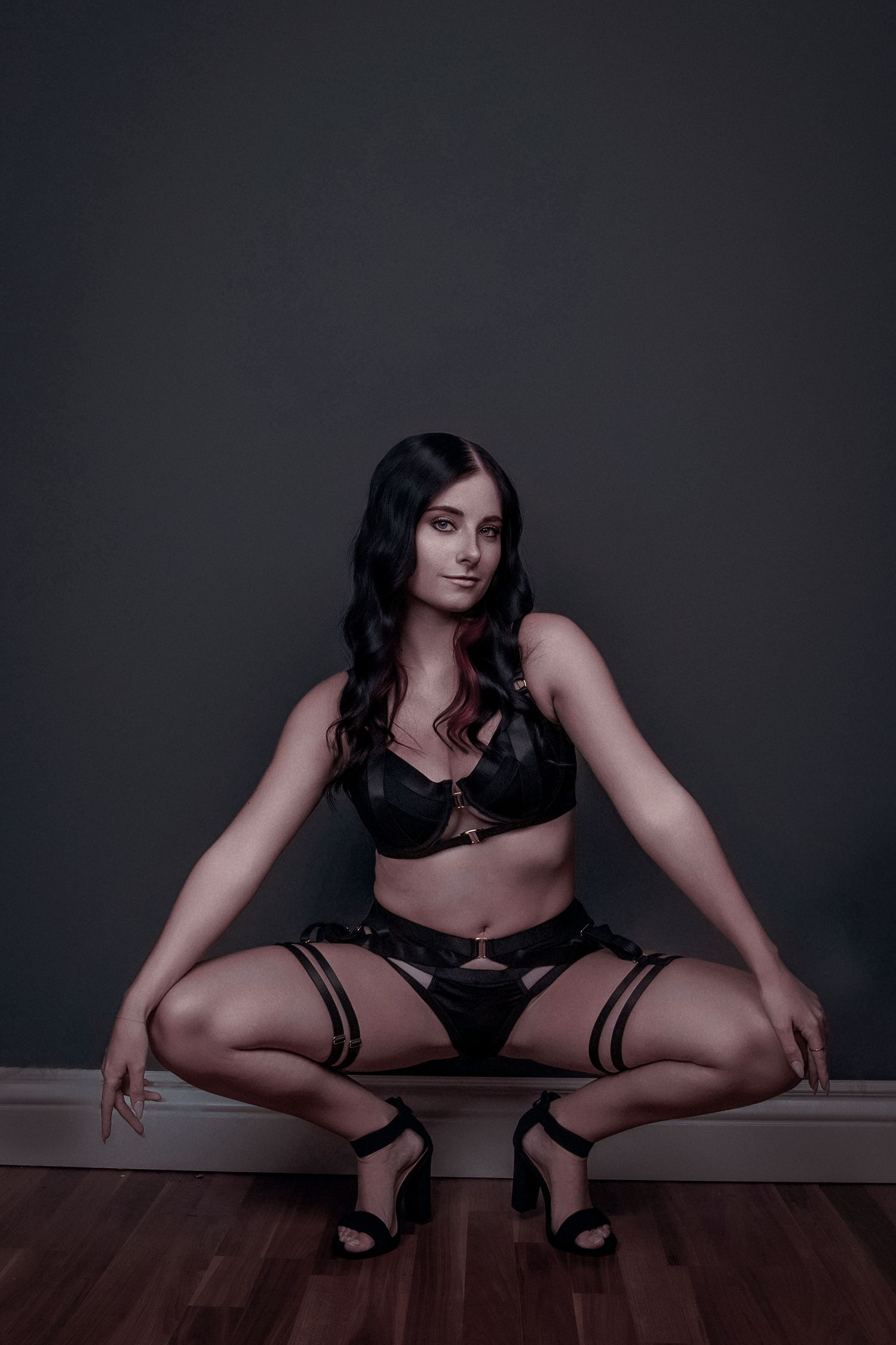 Woman in black lingerie and heels posing confidently in a squatting position against a dark studio background.