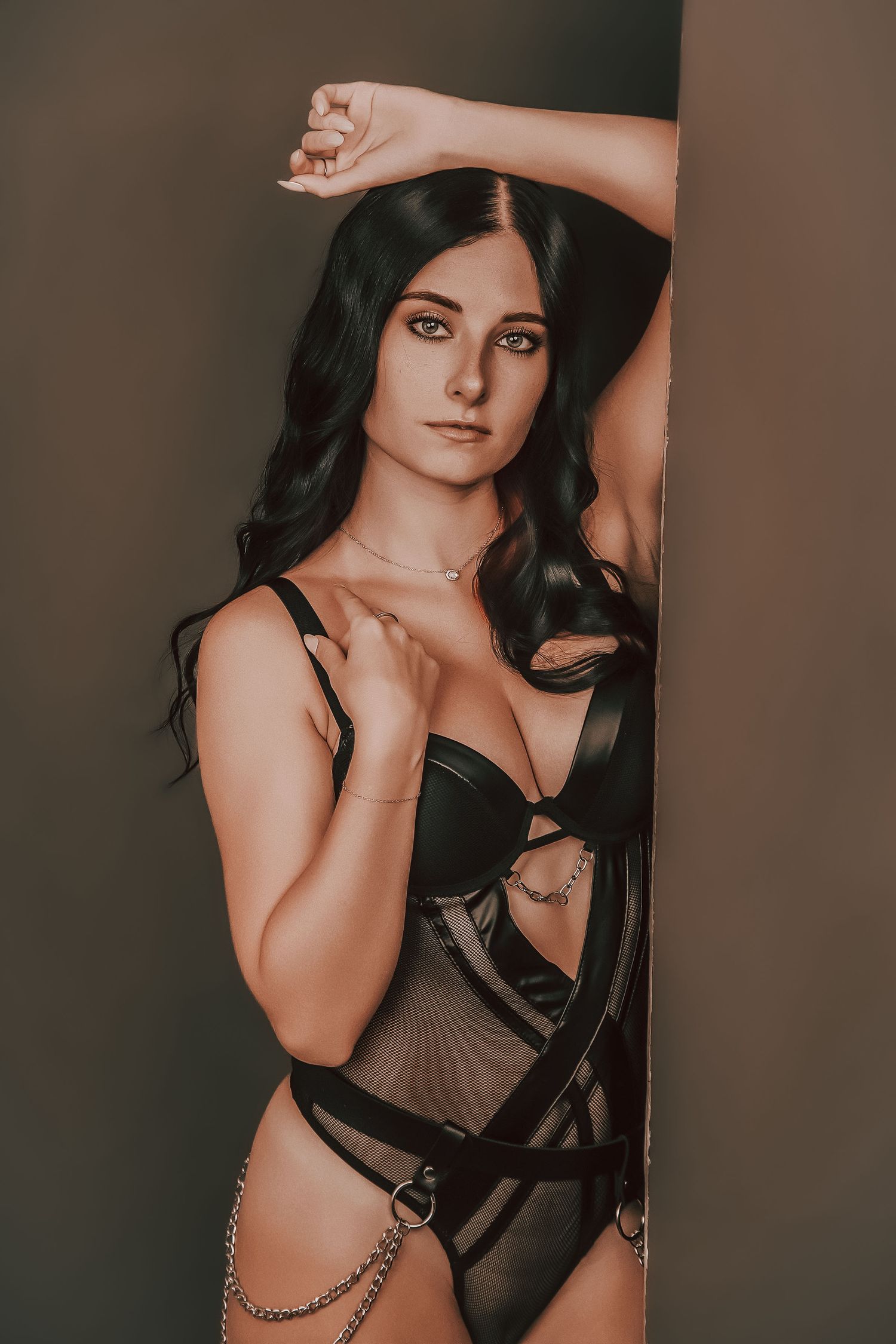 Woman in black lingerie with chain accessories posing confidently against a dark wall.